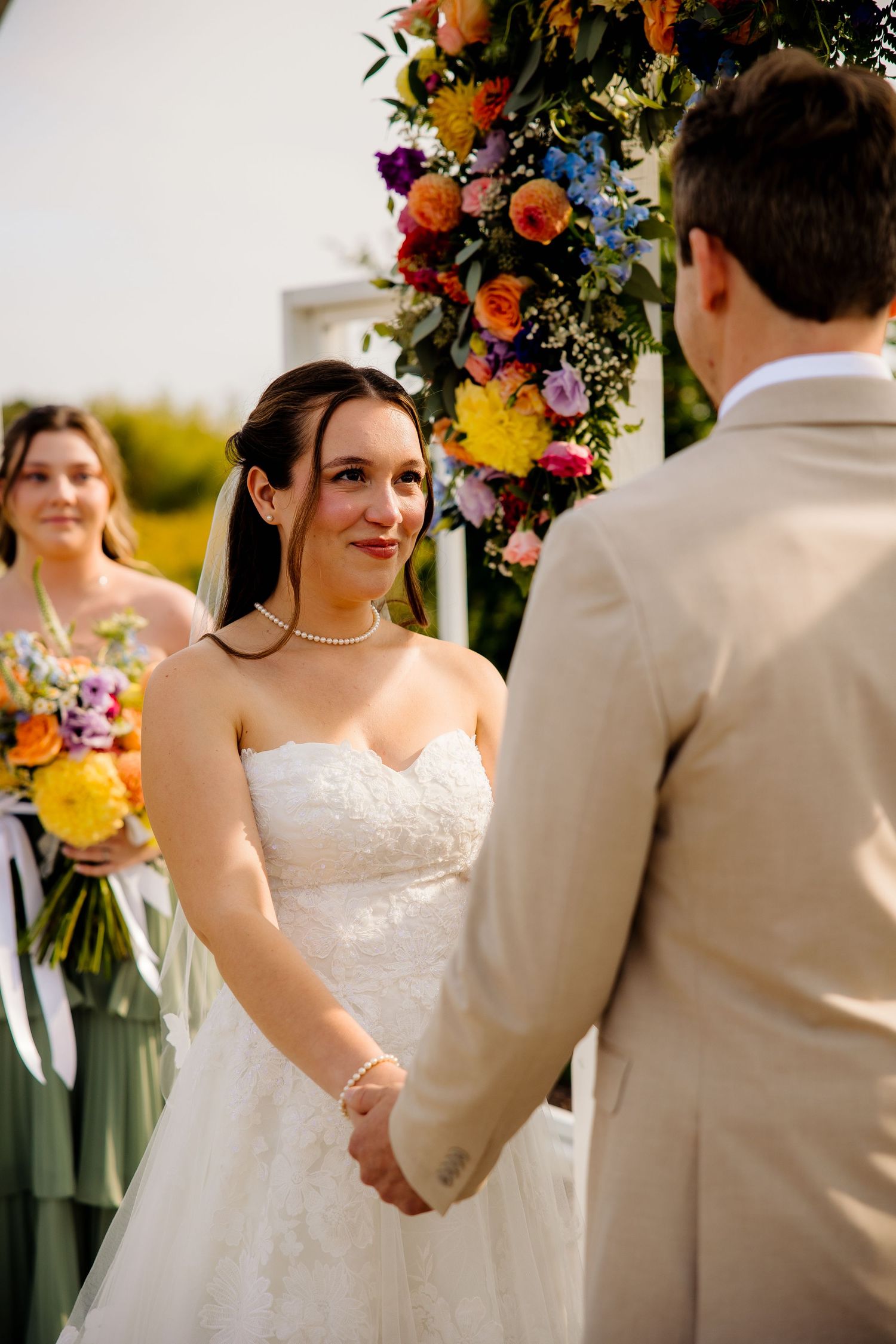 Bride in white strapless gown holds grooms hands at outdoor wedding ceremony with colorful floral arch backdrop.
