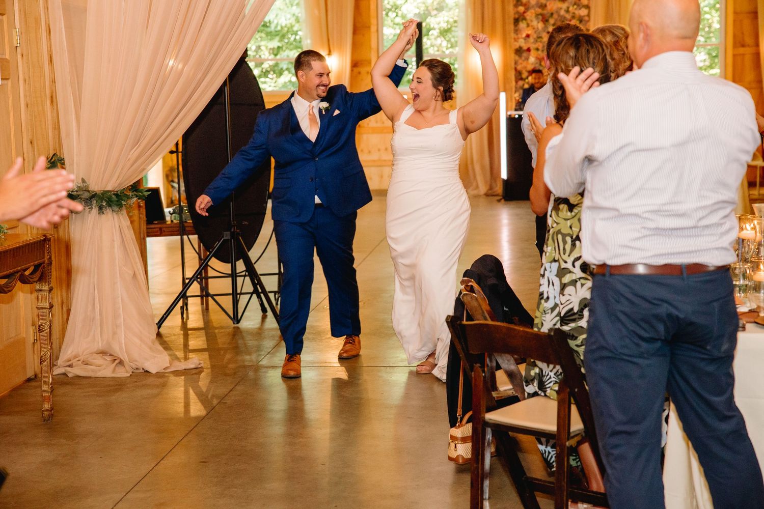 Bride and groom make grand entrance at wedding reception, groom in blue suit, bride in white gown, guests applauding.