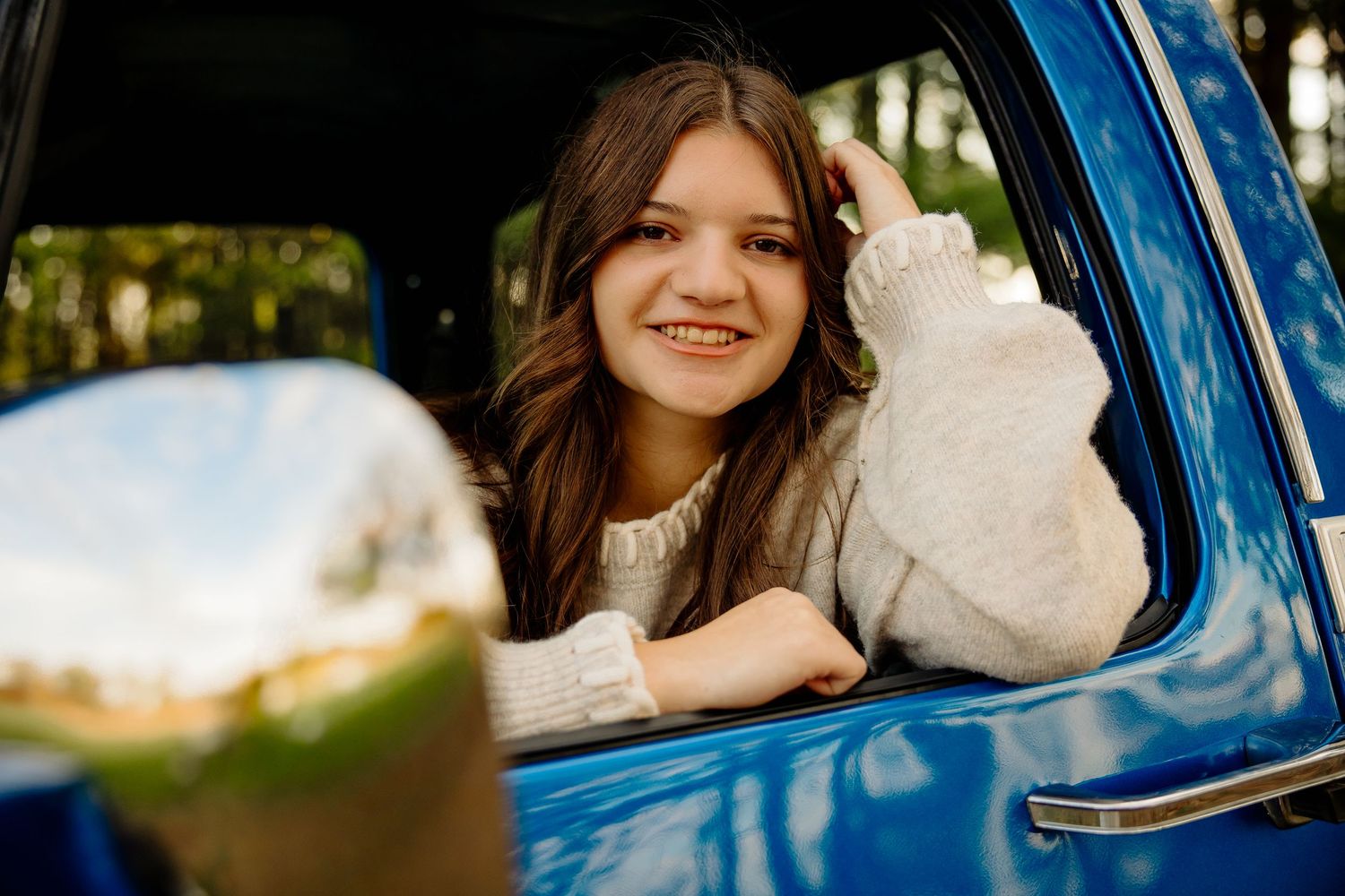 Young woman with long brown hair smiling from a blue vintage truck window, leaning on the door in a cozy sweater.