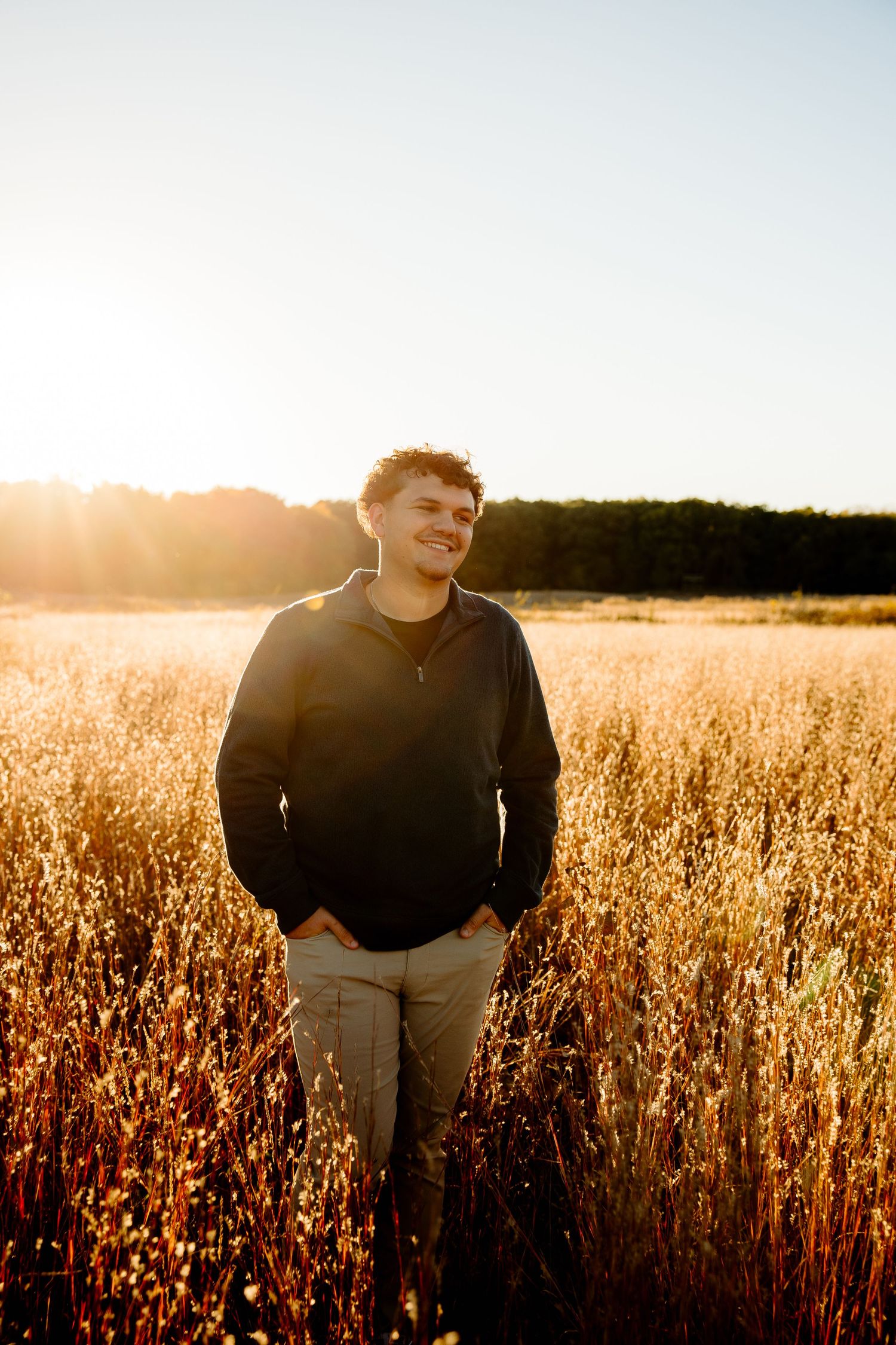 Young man smiling in golden field at sunset, wearing black hoodie and khaki pants, with warm sunlight glowing behind him.