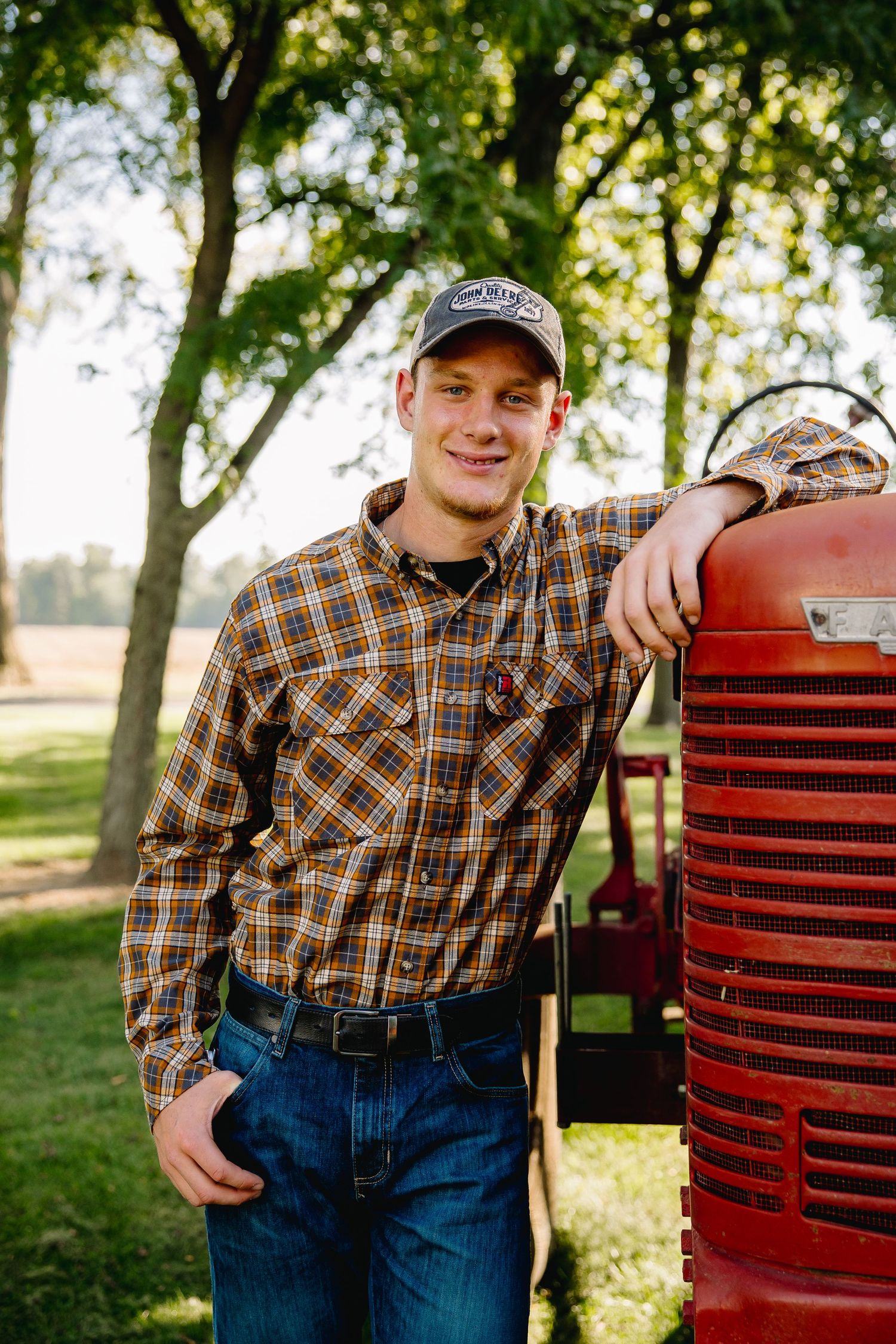 Young man in plaid shirt and cap leans on vintage red tractor surrounded by green trees on a sunny farm day.
