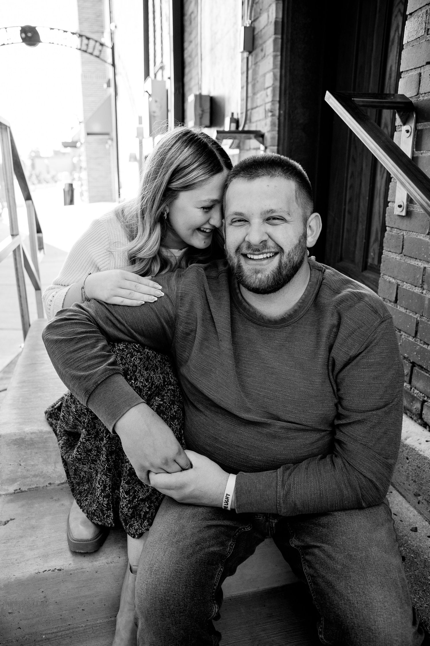 Black and white photo of a laughing couple sitting on steps, woman embracing man from behind in urban setting.