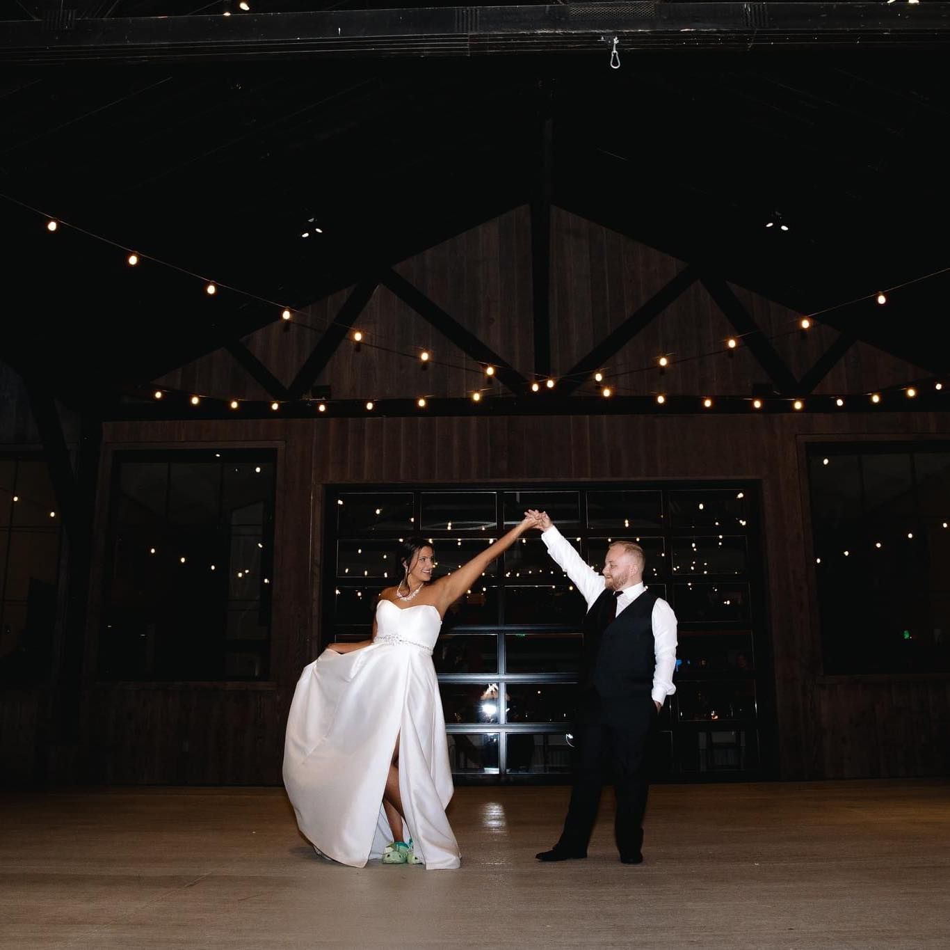Bride and groom share first dance in rustic barn venue with warm string lights overhead.