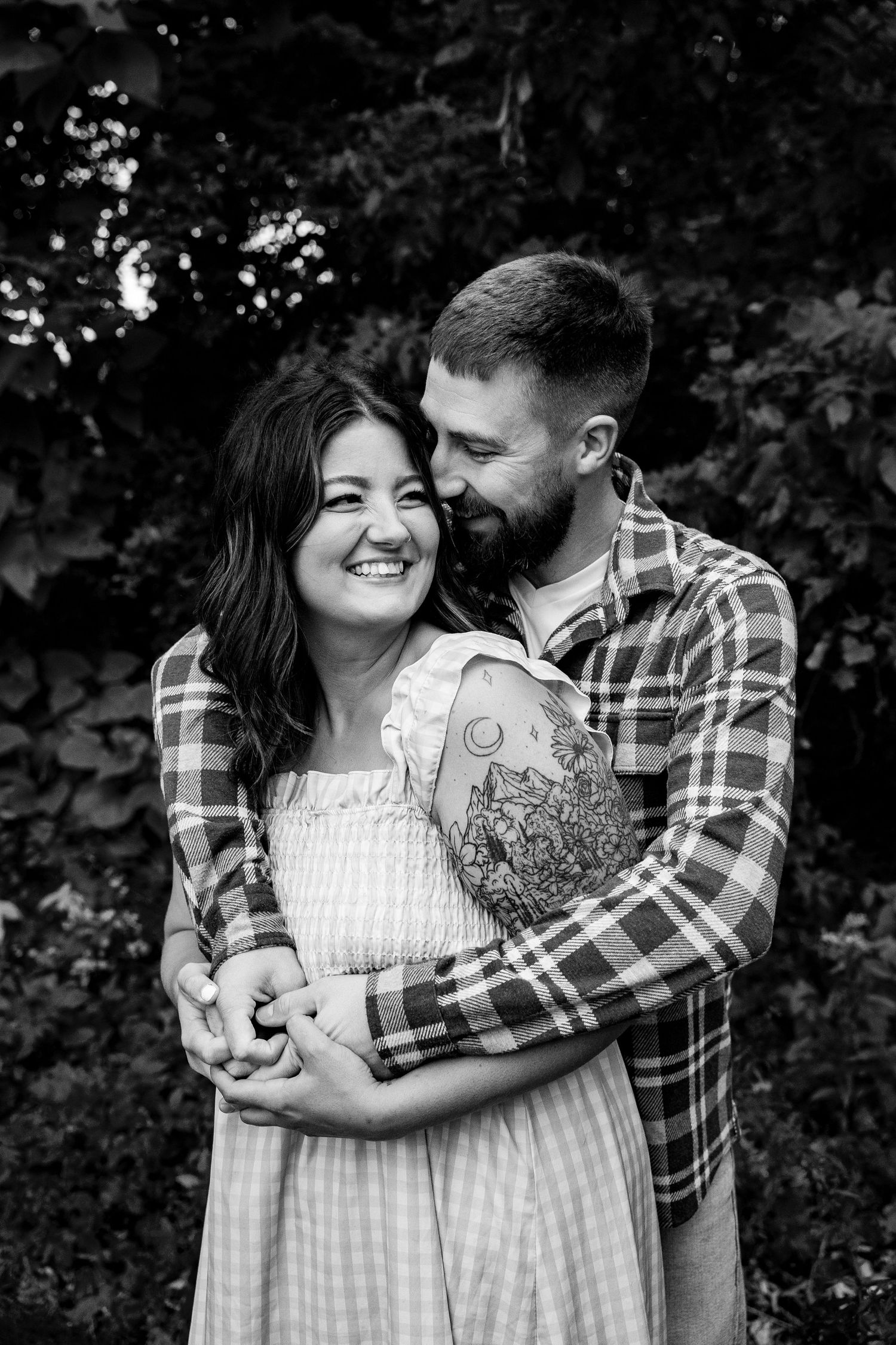Black and white photo of a smiling couple embracing outdoors, man with tattoed arm hugging woman in sundress.