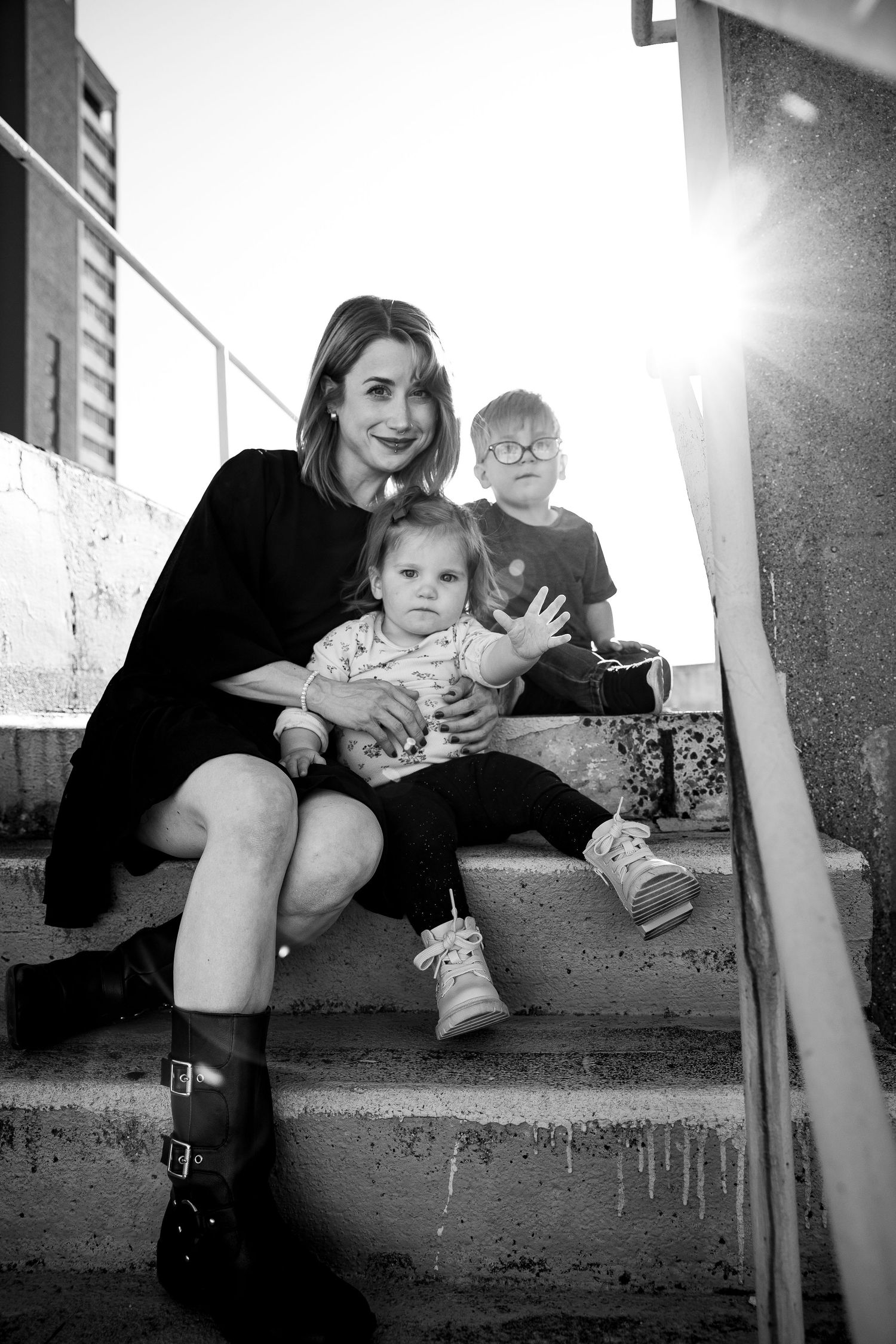 Black and white photo of a woman in black dress sitting on steps with two young children, backlit by sunlight.