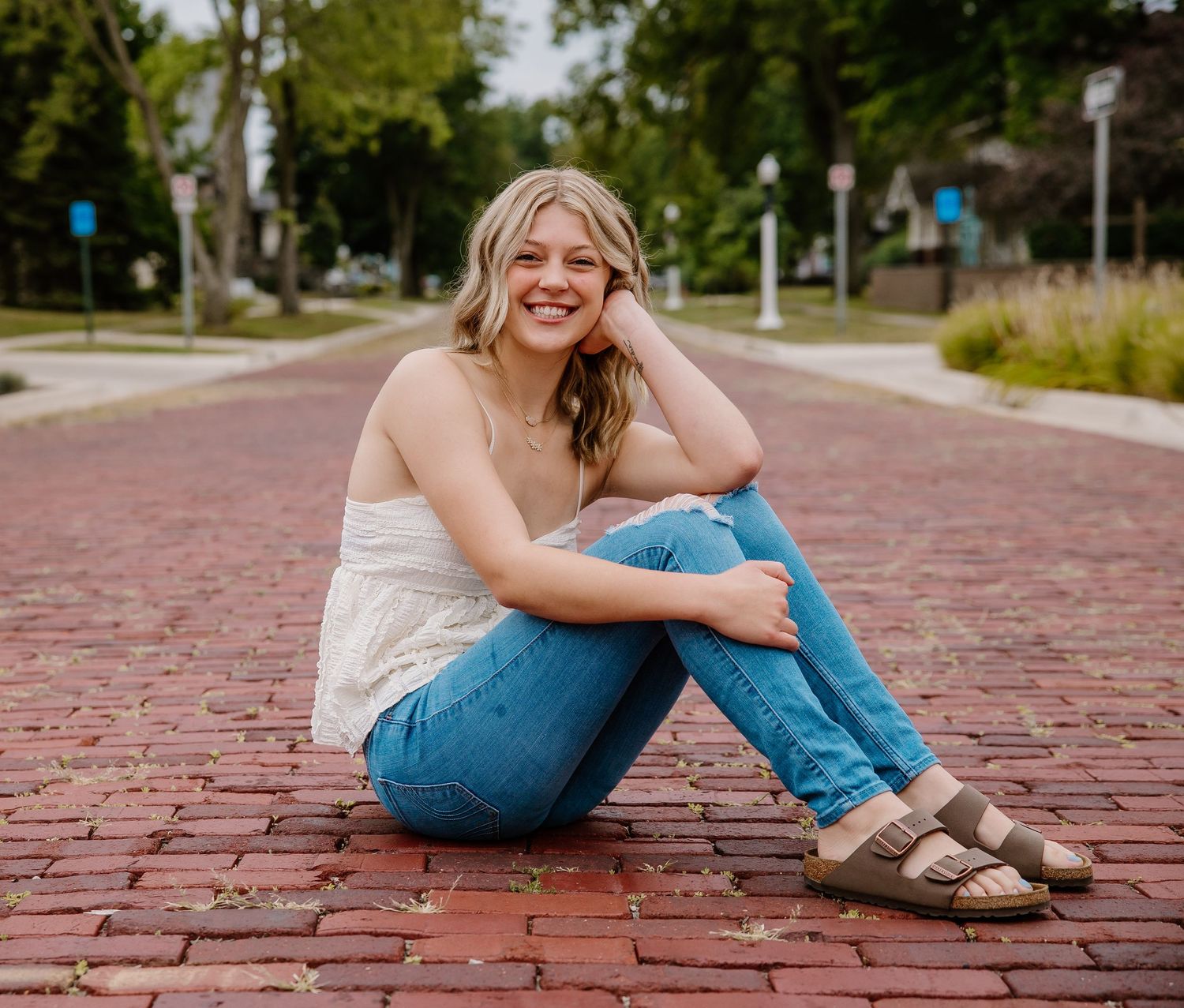 A smiling young woman sits on a red brick path in jeans and white top, posing casually outdoors.