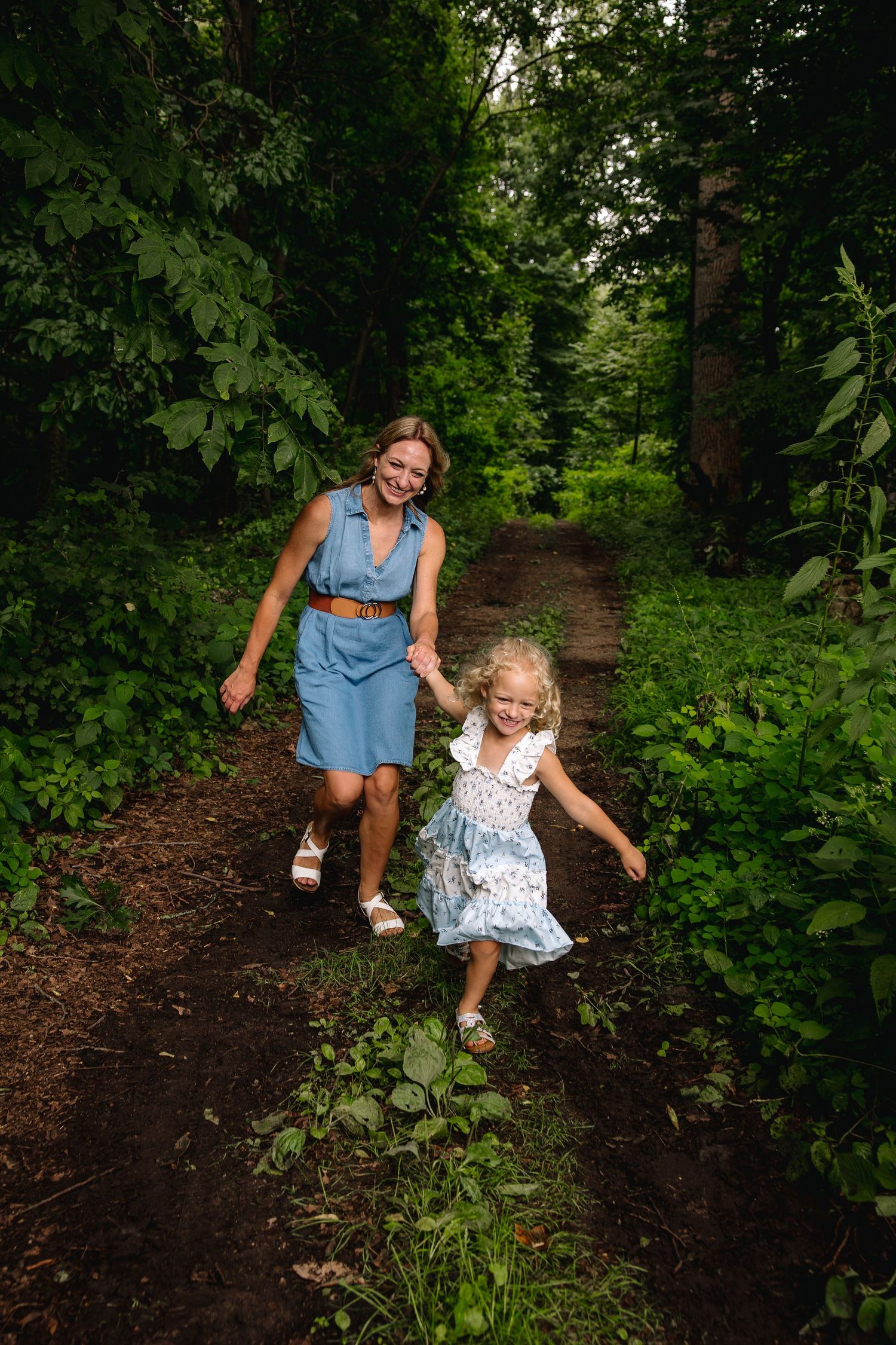 Mother and daughter holding hands walking on a forest path surrounded by lush green trees.
