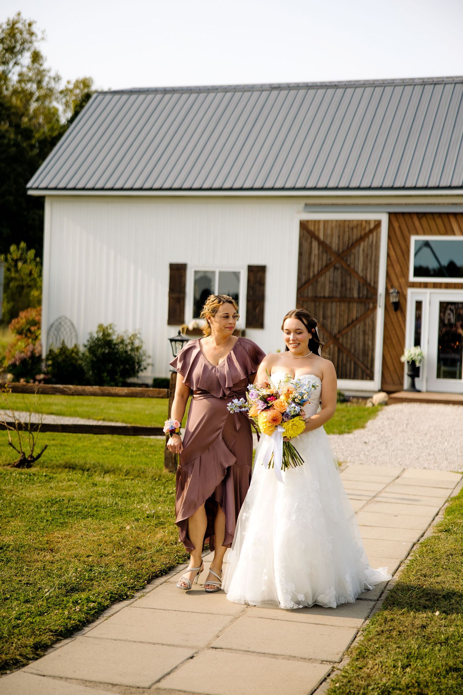 Bride in white gown walks with bridesmaid in mauve dress, holding colorful bouquet outside a white barn venue.