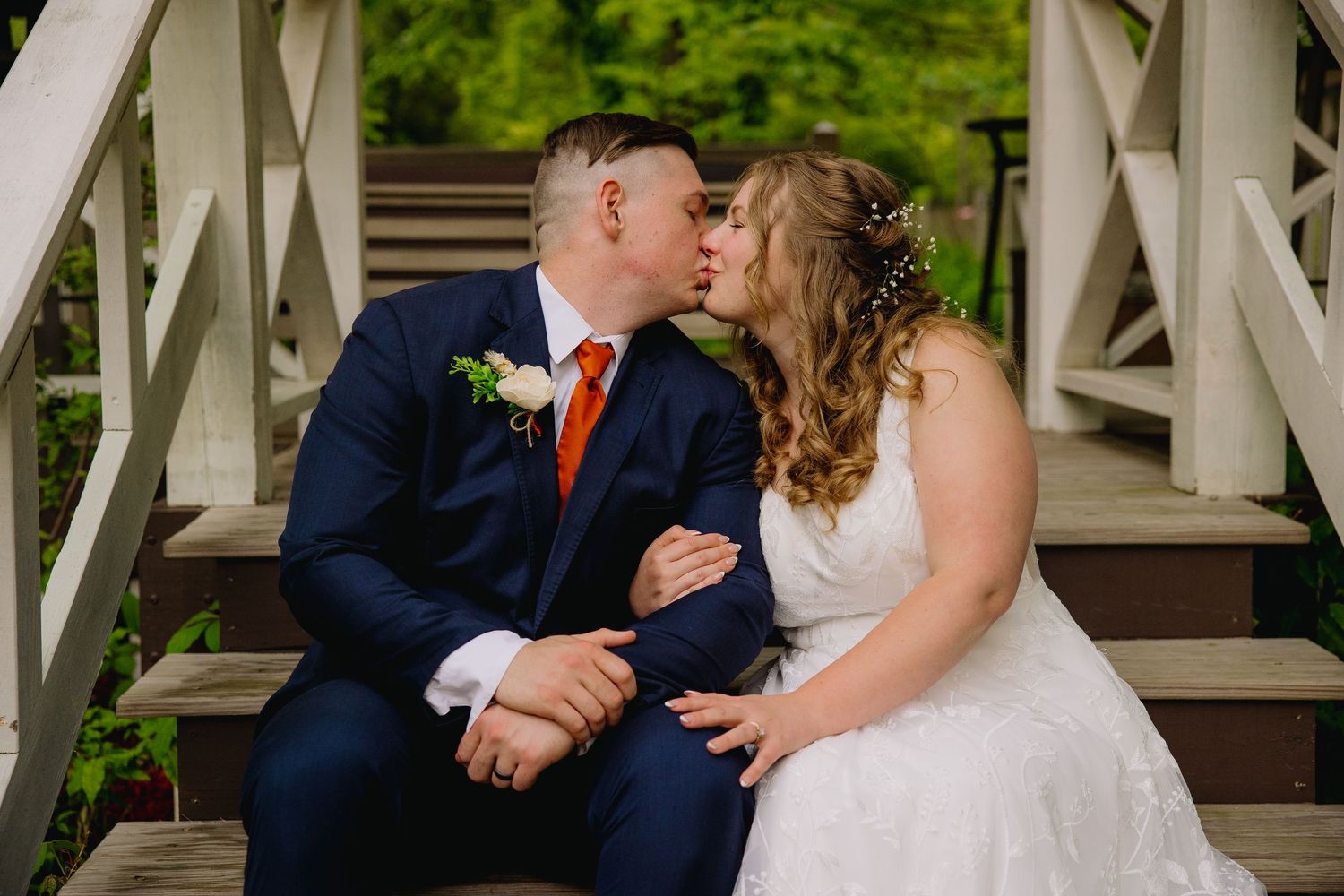 Bride and groom share a romantic kiss on white wooden steps, groom in navy suit with orange tie, bride in white lace gown.