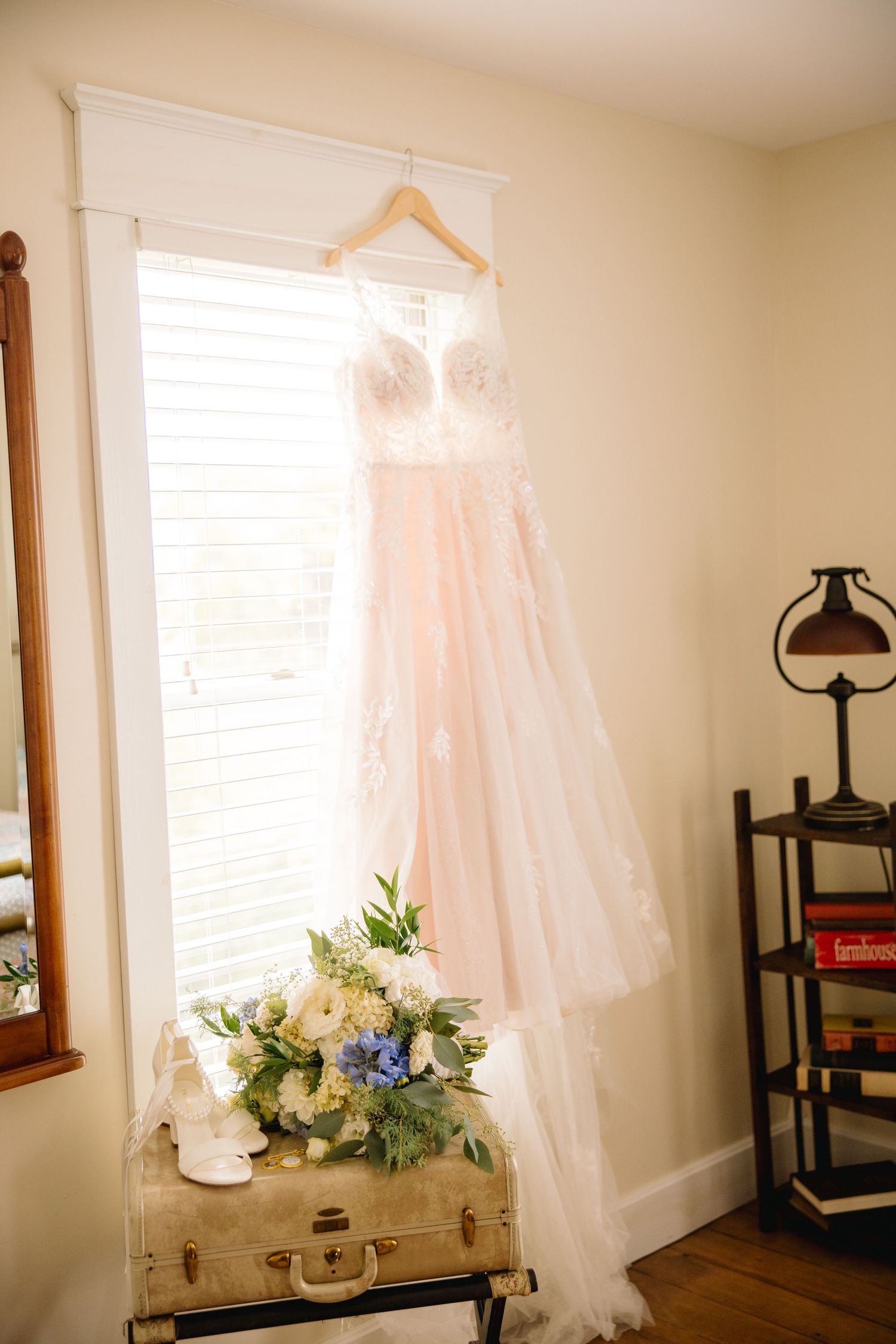 Elegant blush wedding dress hanging in window above vintage suitcase with bridal bouquet and white heels.