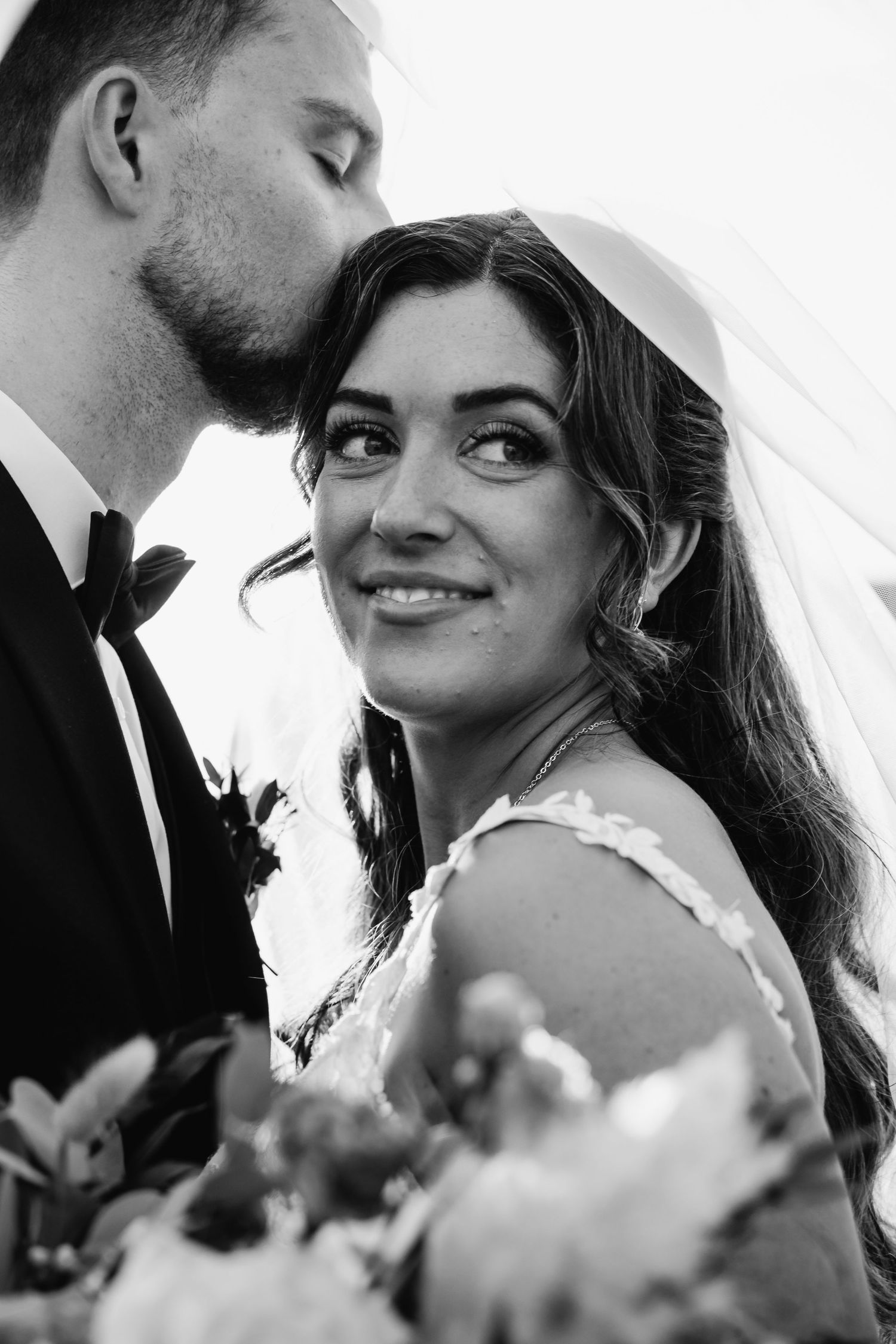 Black and white wedding photo of smiling bride with veil, groom kissing her head, holding bouquet.