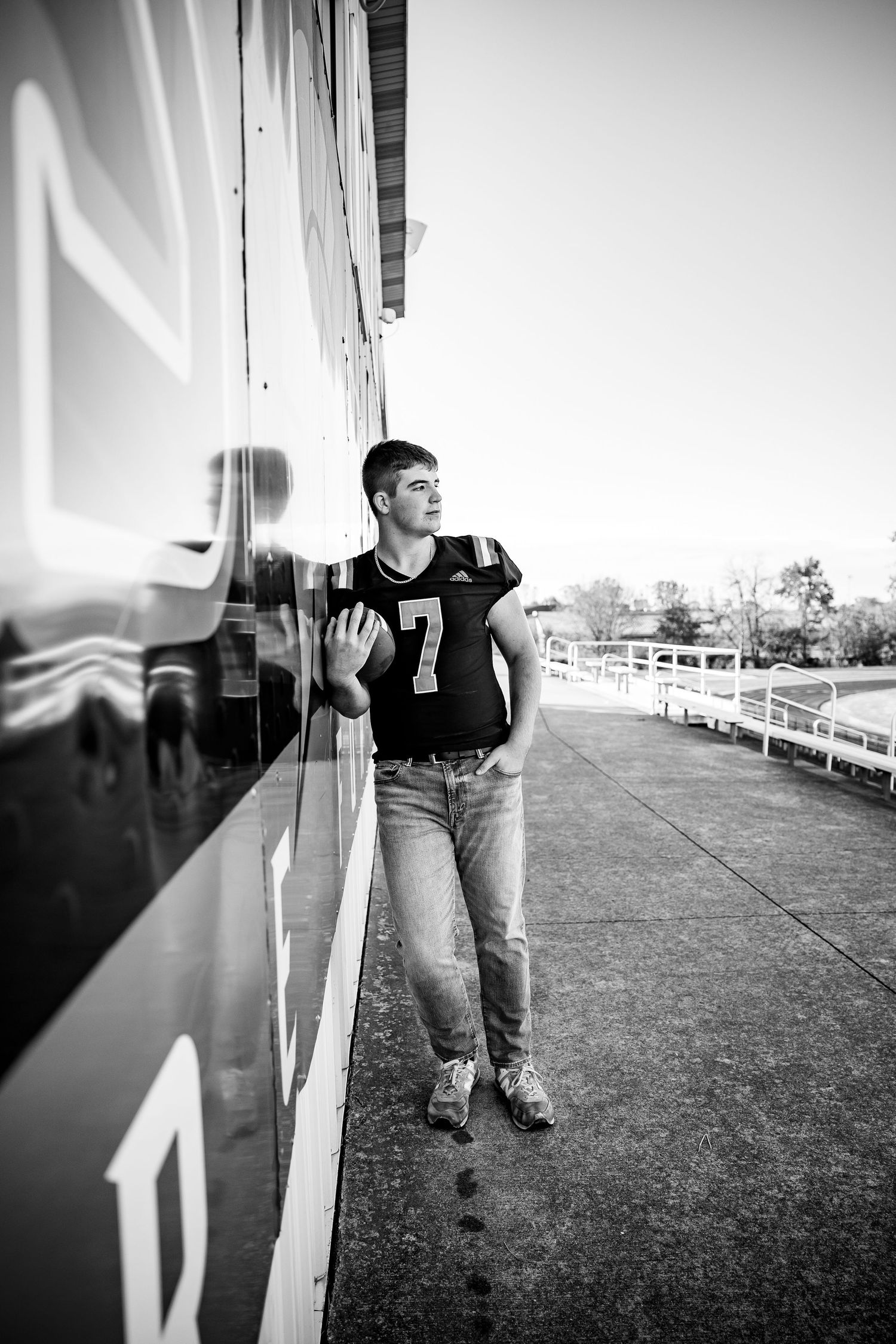 Black and white photo of a teen boy in football jersey number 7 leaning against a stadium wall in jeans.