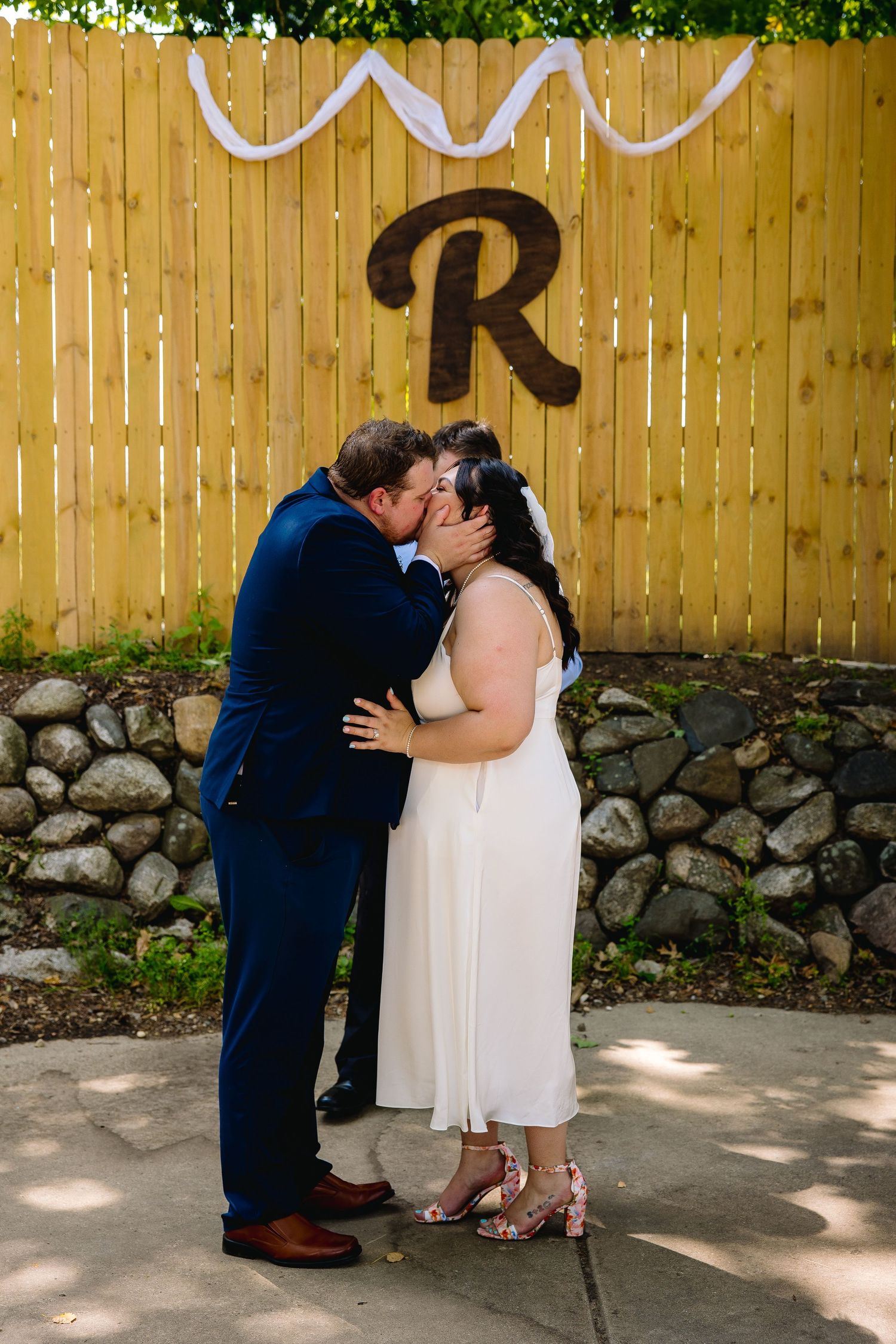 Bride and groom share first kiss in front of wooden fence decorated with letter R monogram at outdoor wedding ceremony.