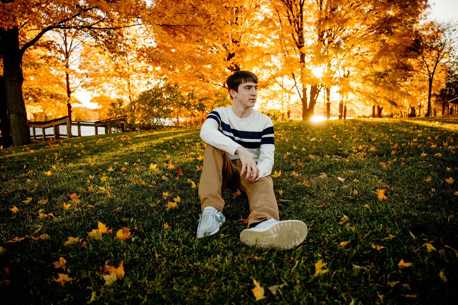 Young man sitting on grass surrounded by vibrant orange autumn foliage during golden hour in a park.