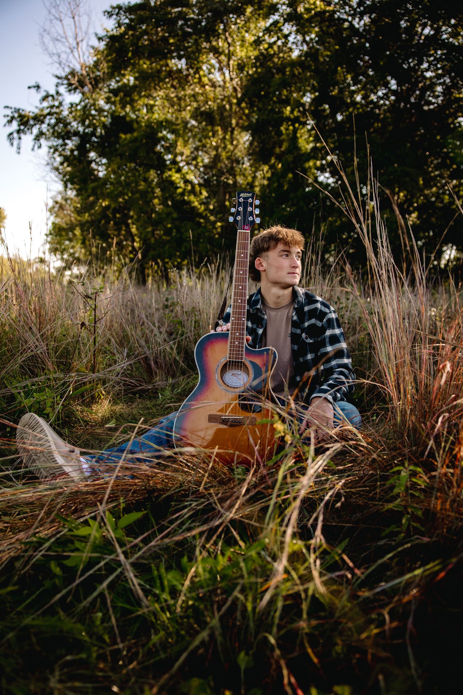 Young man sitting in tall grass at golden hour, leaning against an acoustic guitar, wearing a flannel shirt outdoors.