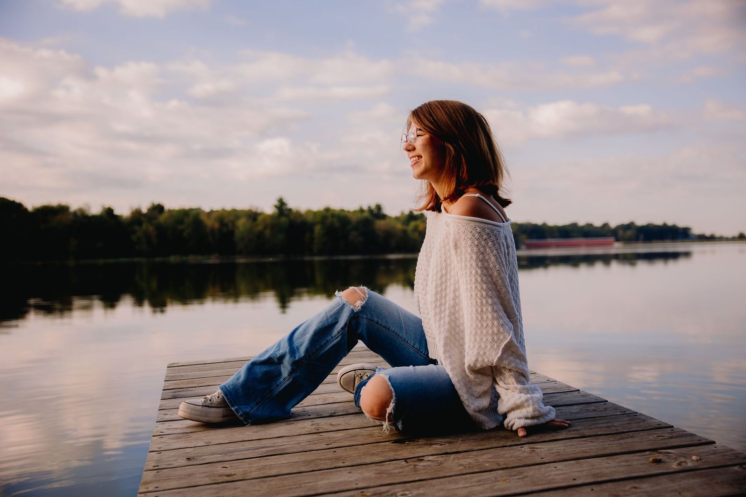 Young woman in ripped jeans and white sweater sitting on wooden dock by calm lake at sunset.