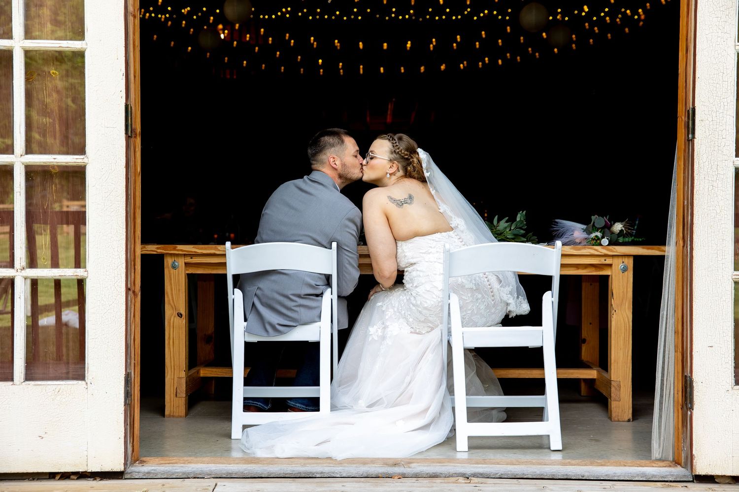 Newlywed couple sharing a romantic kiss at their wedding reception table, bride in white gown with veil, groom in gray suit.
