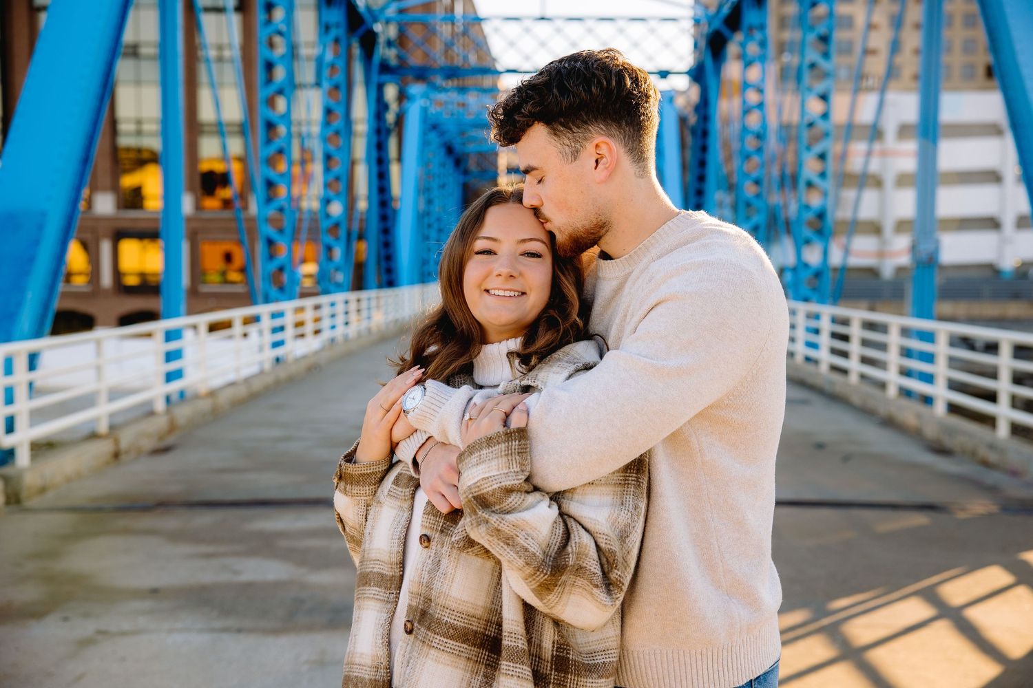 Couple embracing on a blue steel bridge, woman smiling in plaid jacket, man kissing her head, golden hour lighting.