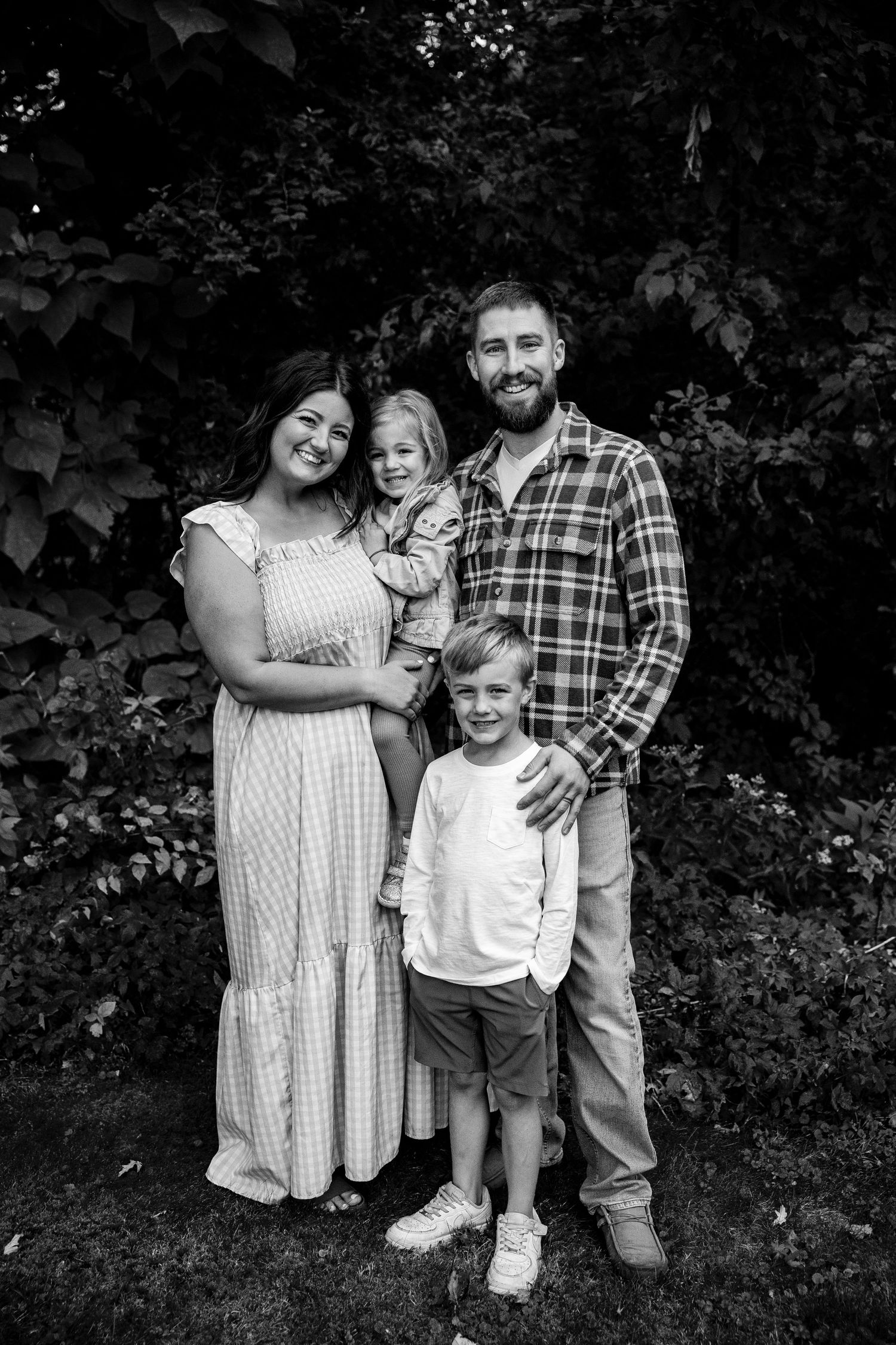 Black and white family portrait of parents with two young children standing in front of lush greenery.
