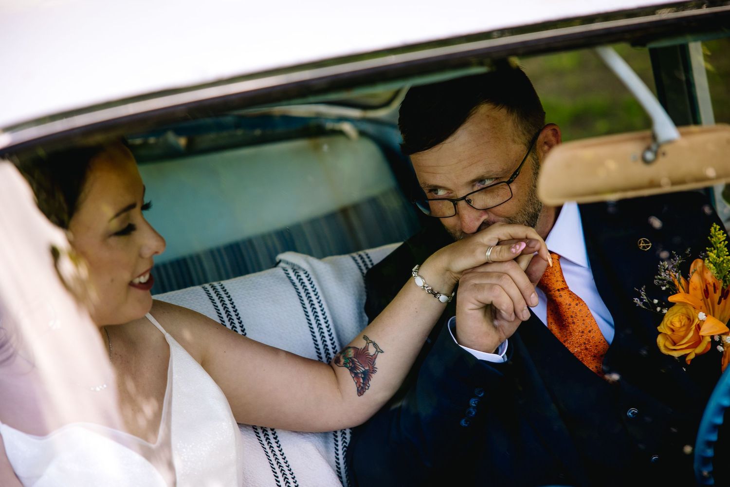 Groom placing wedding ring on bride's finger in vintage car, bride wearing veil with floral tattoo visible.