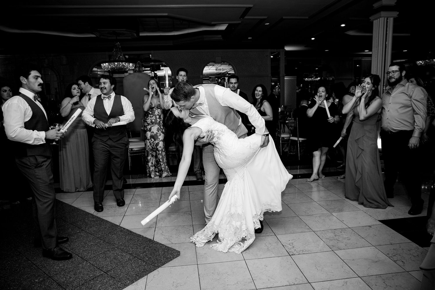 Black and white photo of bride and groom sharing a dramatic dip dance surrounded by cheering wedding guests.