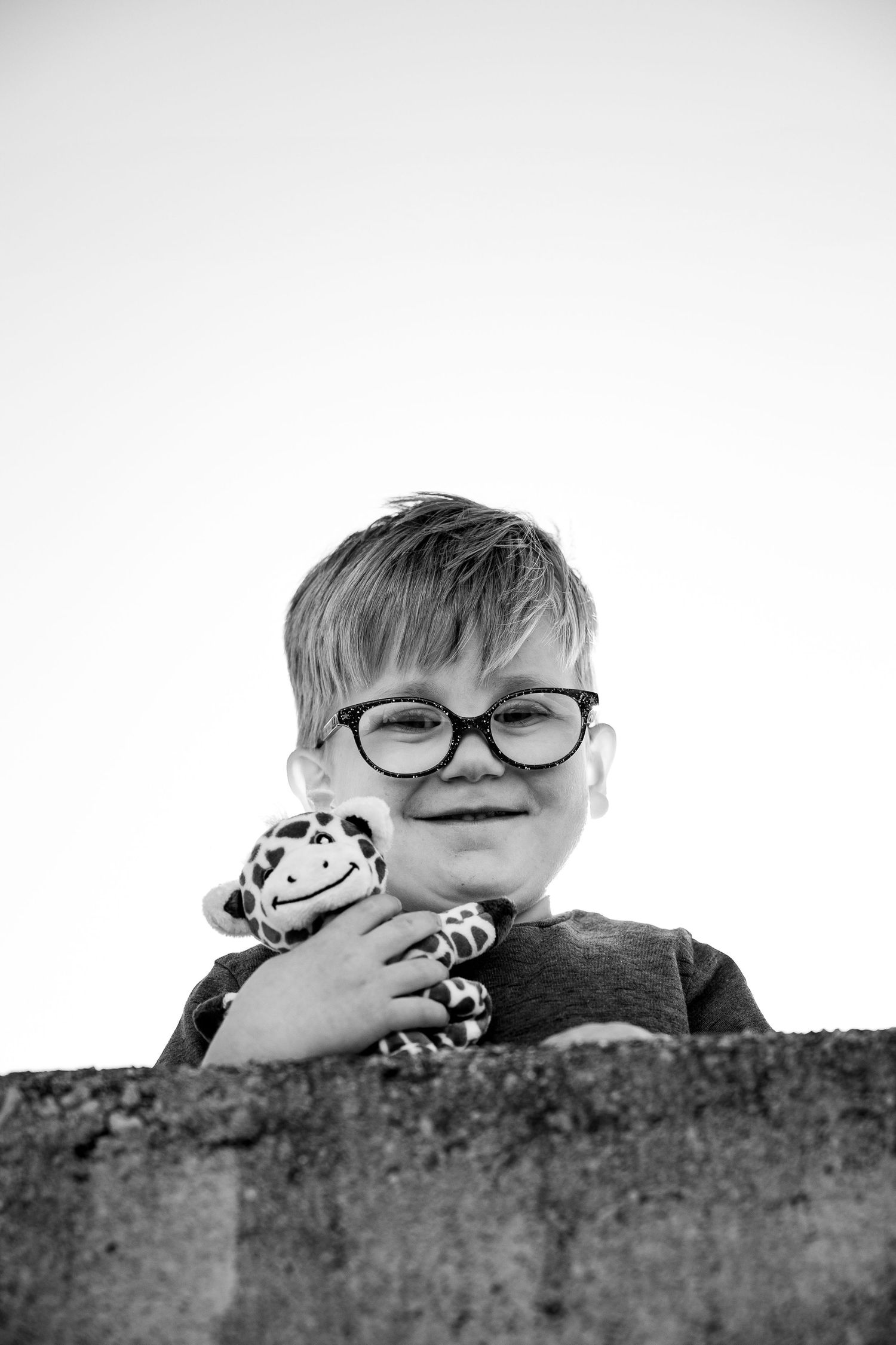 Black and white photo of a smiling boy with glasses hugging a stuffed animal toy while peeking over a stone wall.