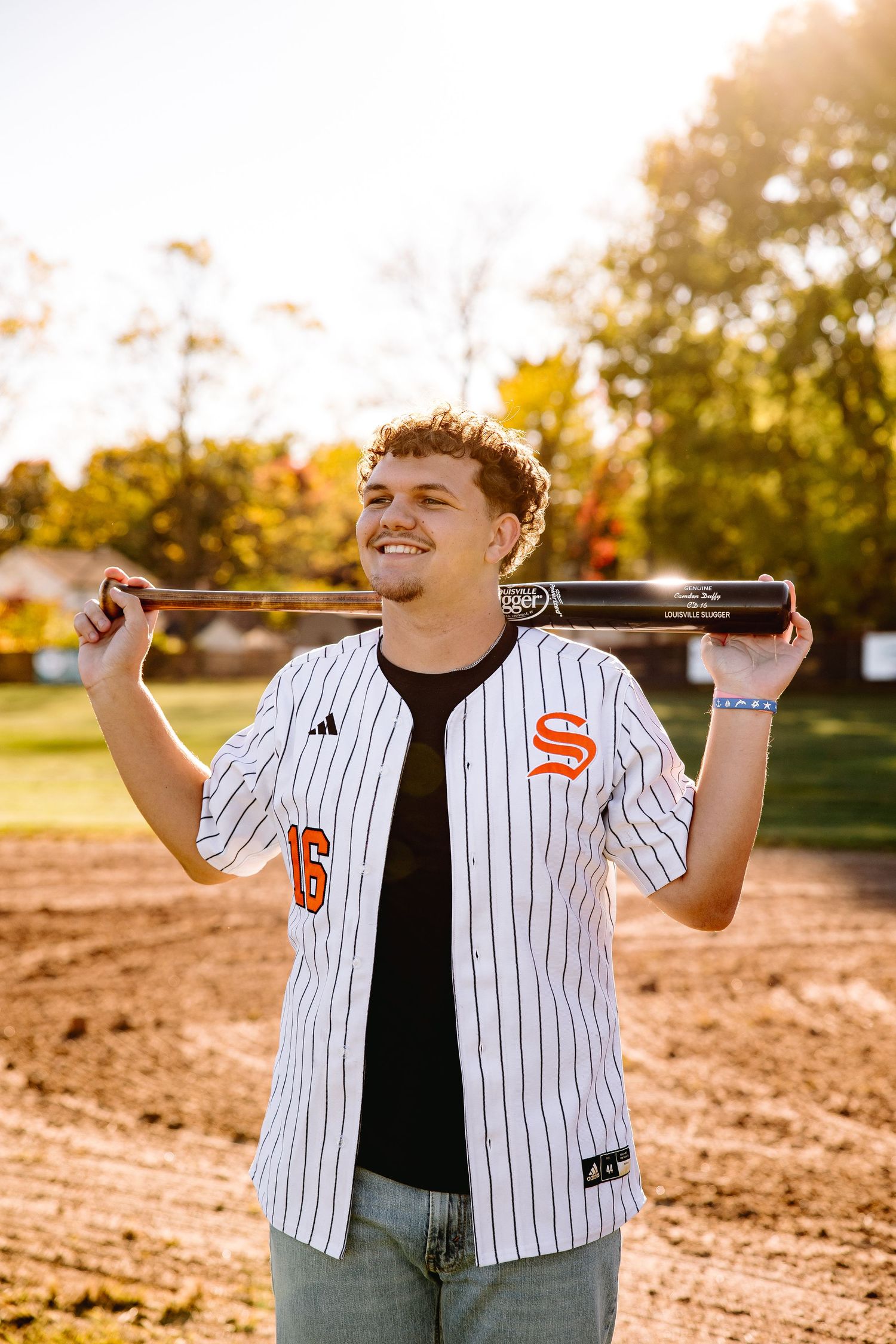 Smiling young man in white pinstripe baseball jersey holding bat behind shoulders on sunny baseball field.