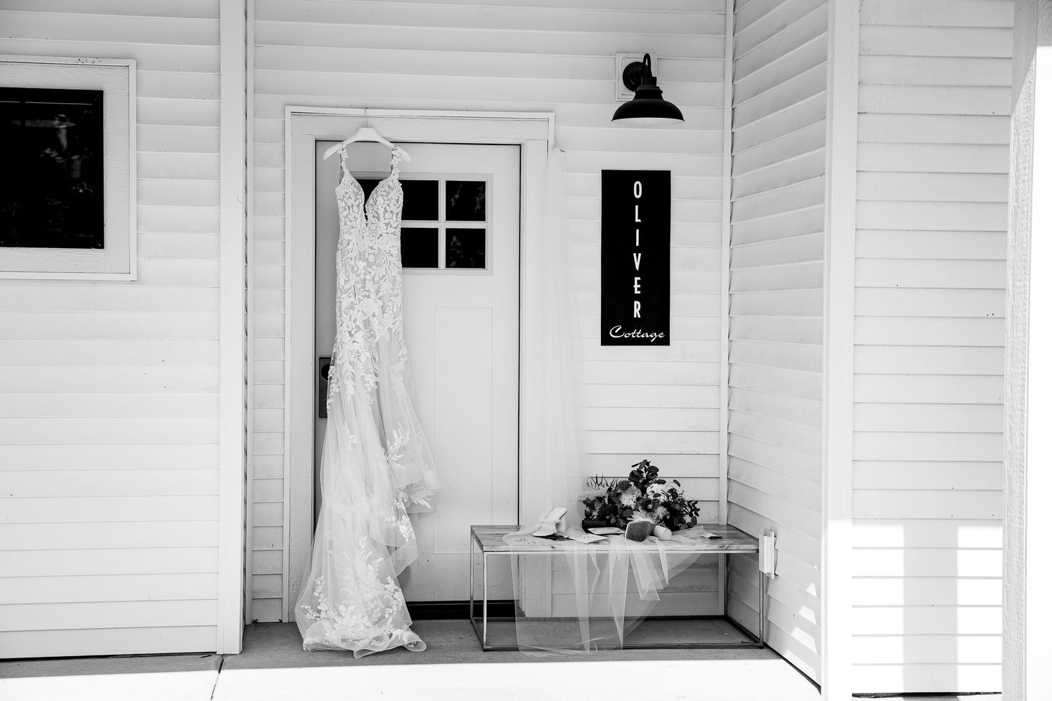 Black and white photo of a lace wedding dress hanging on a white cottage door with bridal accessories on a bench.