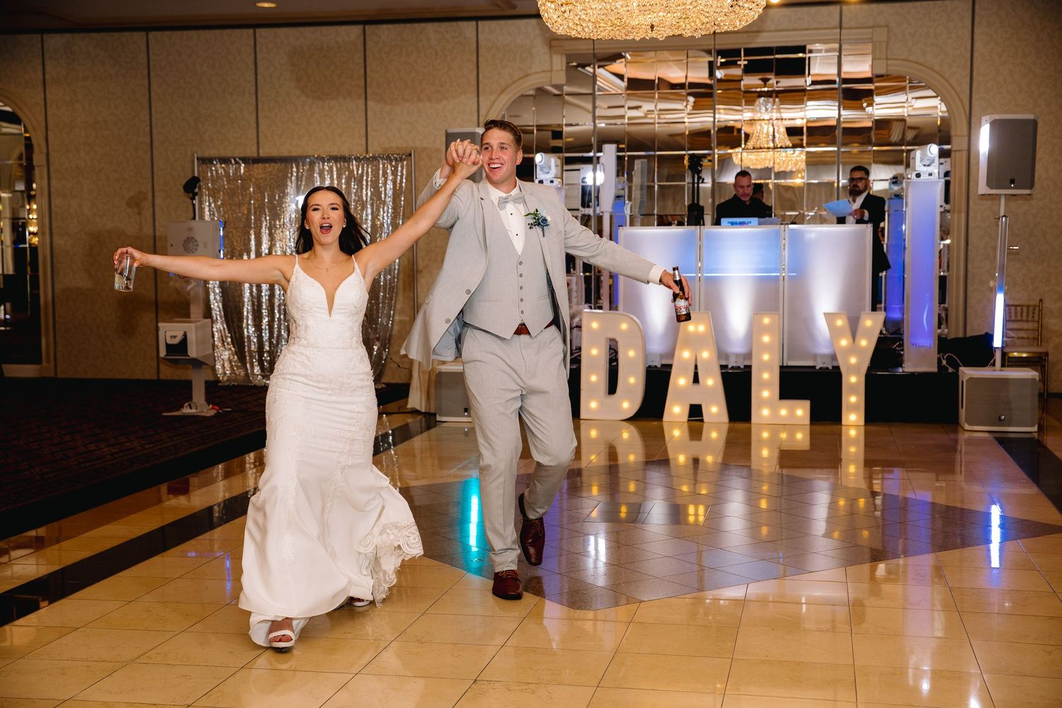 Bride and groom joyfully entering wedding reception with marquee letter DALY sign on dance floor.