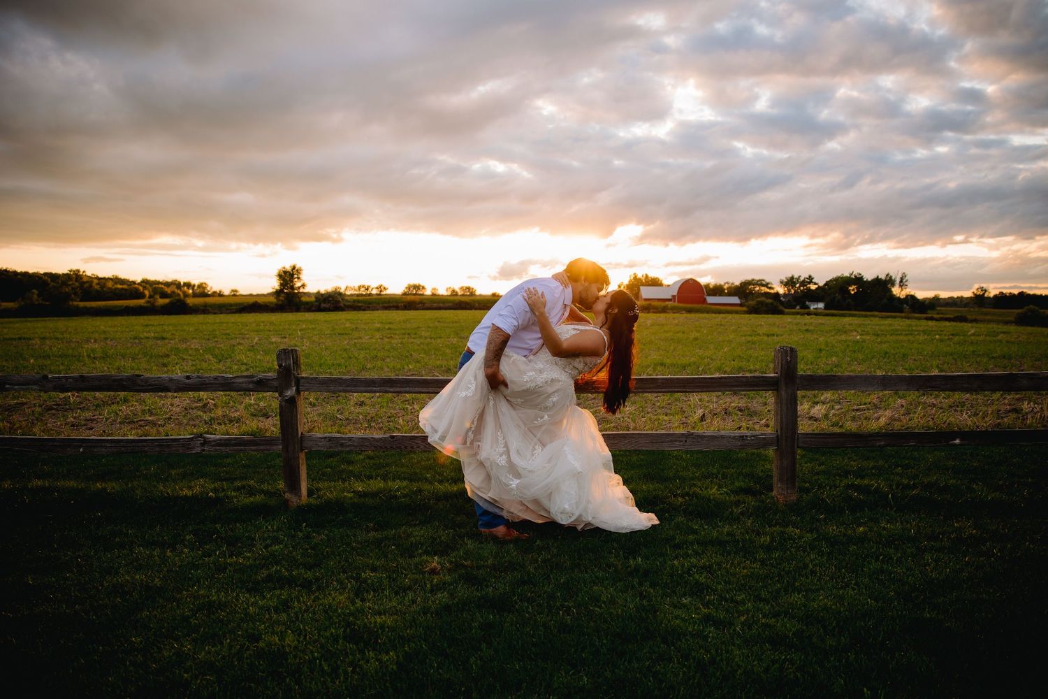 Bride and groom share romantic dip kiss at sunset by rustic fence in green field.