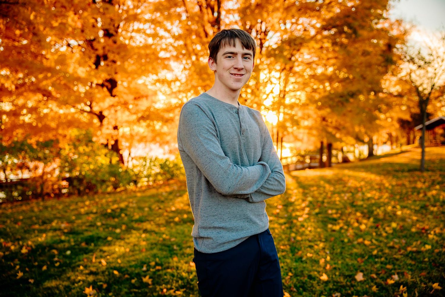 Young man smiling with arms crossed in a vibrant autumn park, surrounded by golden orange foliage trees.
