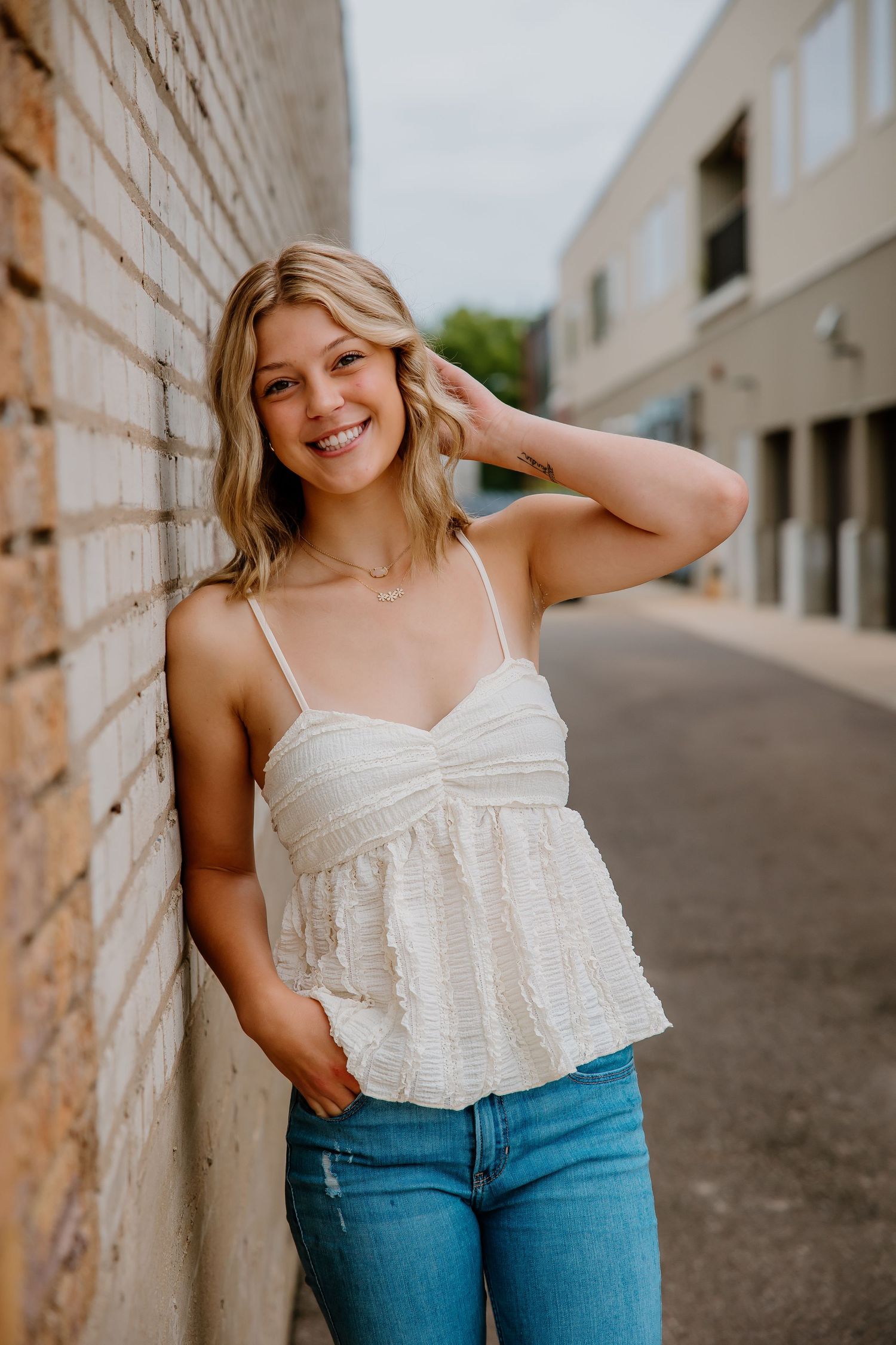 Smiling young blonde woman in white camisole top and jeans leaning against brick wall outdoors.