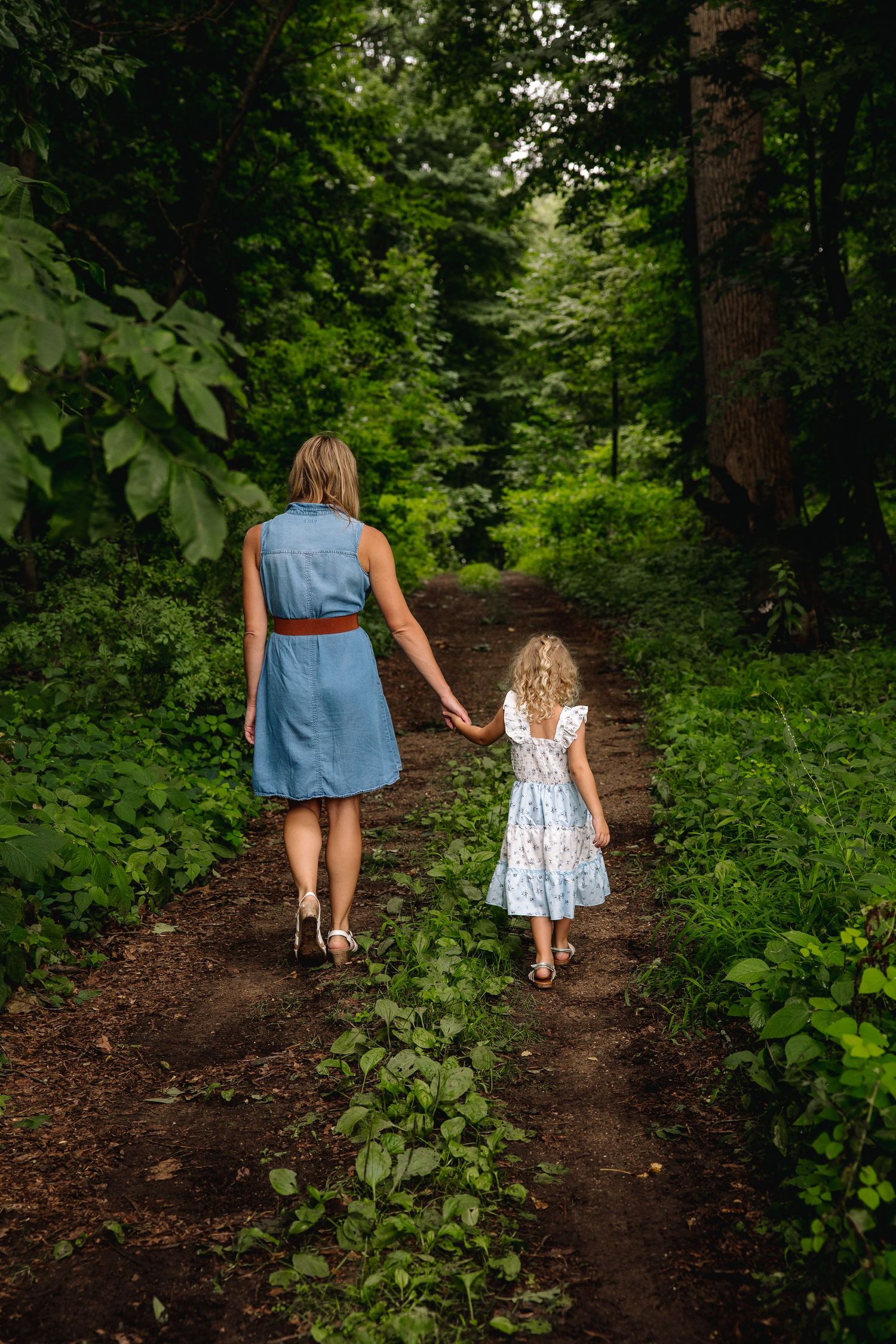 A mother and young daughter walk hand-in-hand along a lush green forest path, both wearing summer dresses.