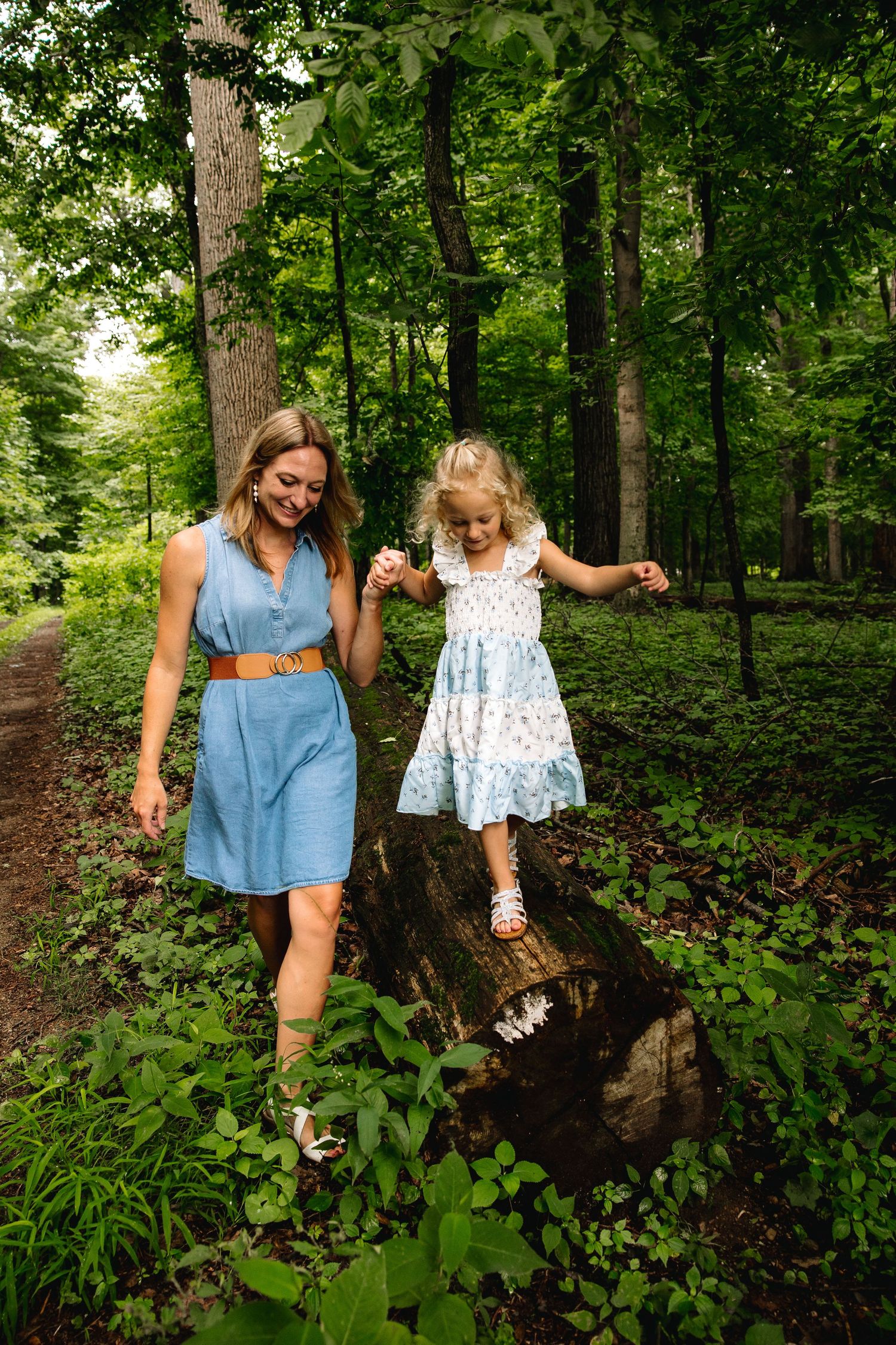 Woman in blue dress holds young girl's hand as she balances on a log in a lush green forest.