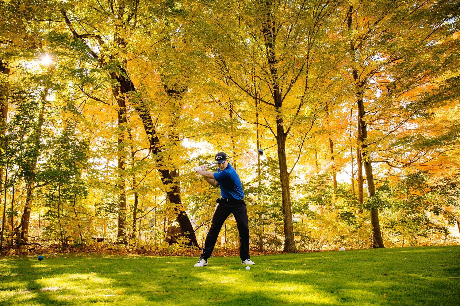 Golfer swinging club on lush green fairway surrounded by vibrant autumn yellow trees in a scenic woodland golf course.