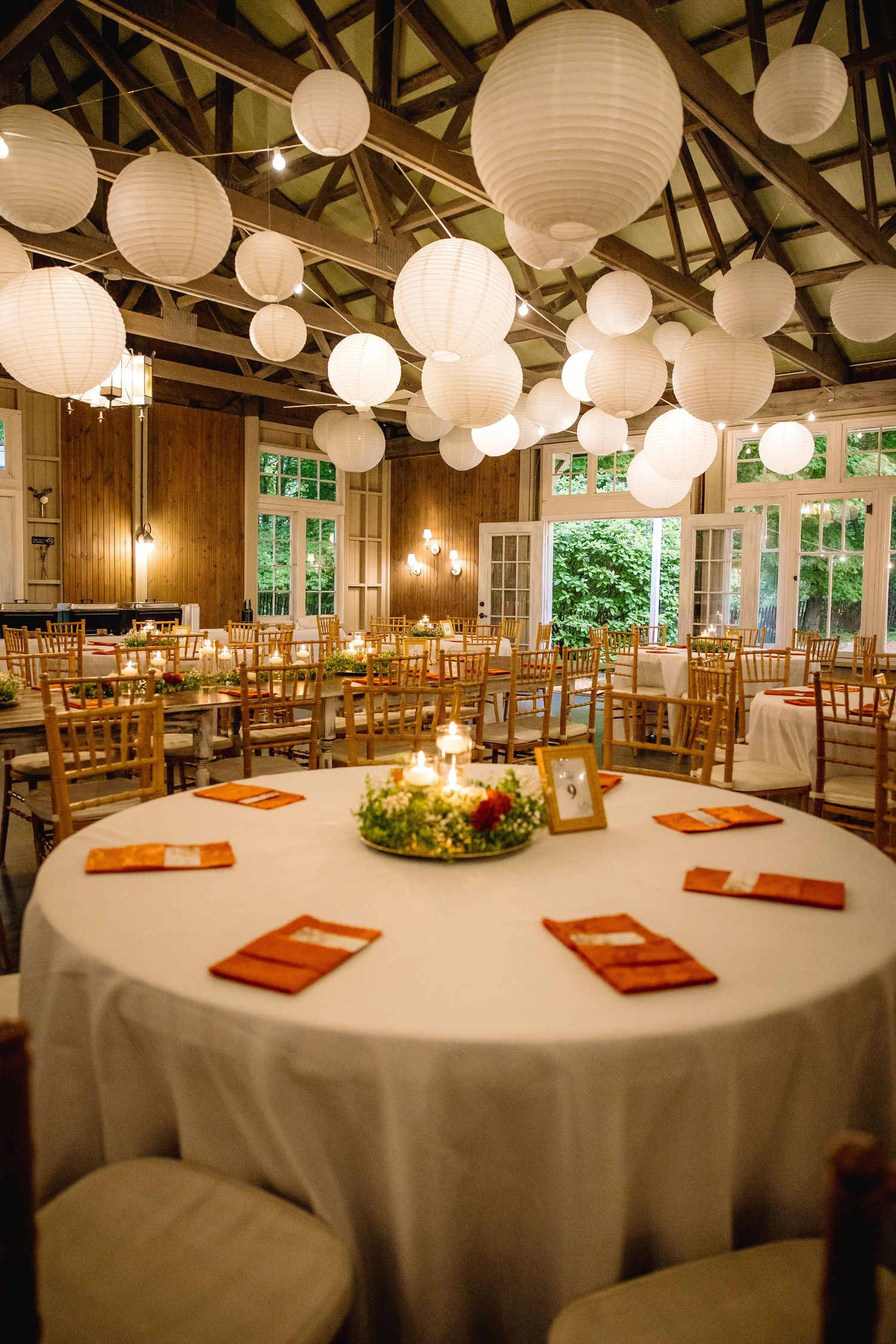 Elegant wedding reception hall with white paper lantern ceiling, round tables, orange napkins, and warm candlelight centerpieces.