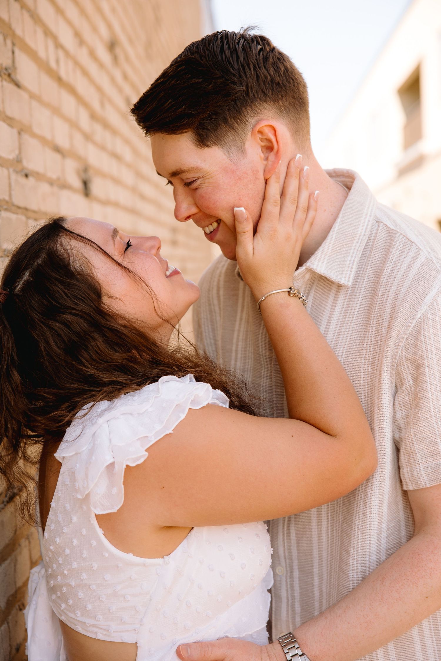 A smiling couple shares a romantic moment against a brick wall, with the woman tenderly holding the man's face.