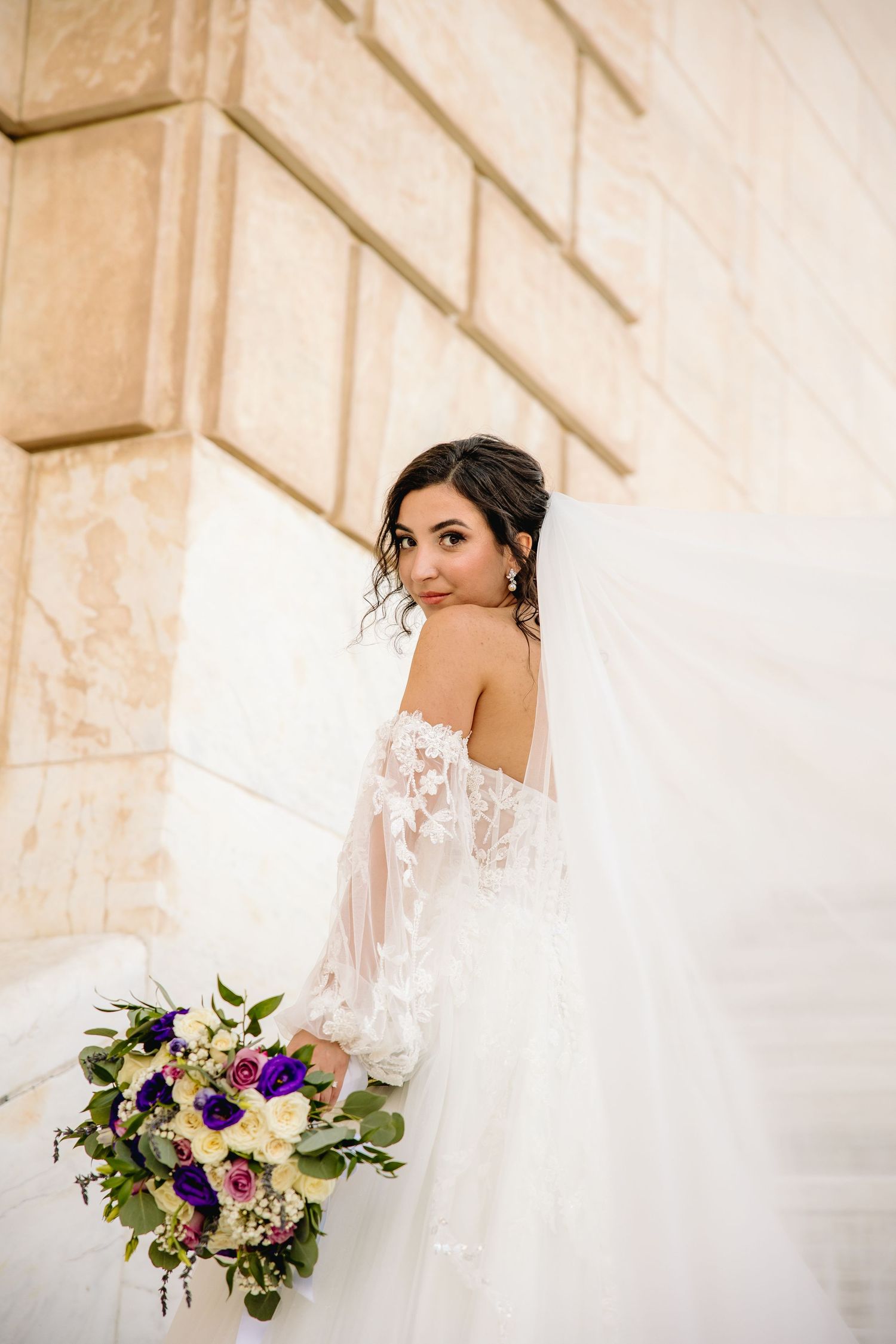 Bride in off-shoulder lace wedding dress holding purple and white bouquet against stone wall architecture.