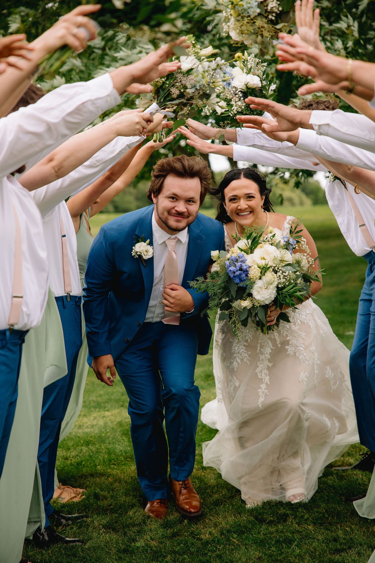 Bride and groom walk through arch of raised hands formed by wedding party outdoors, bride holding white and blue bouquet.