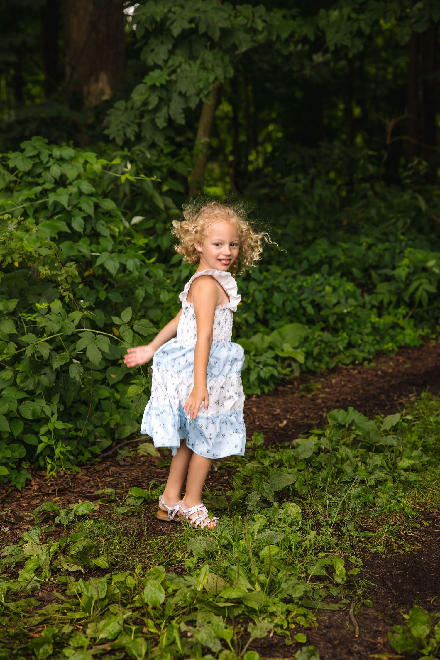 Young girl with curly blonde hair in a blue floral dress walking on a forest path surrounded by green foliage.