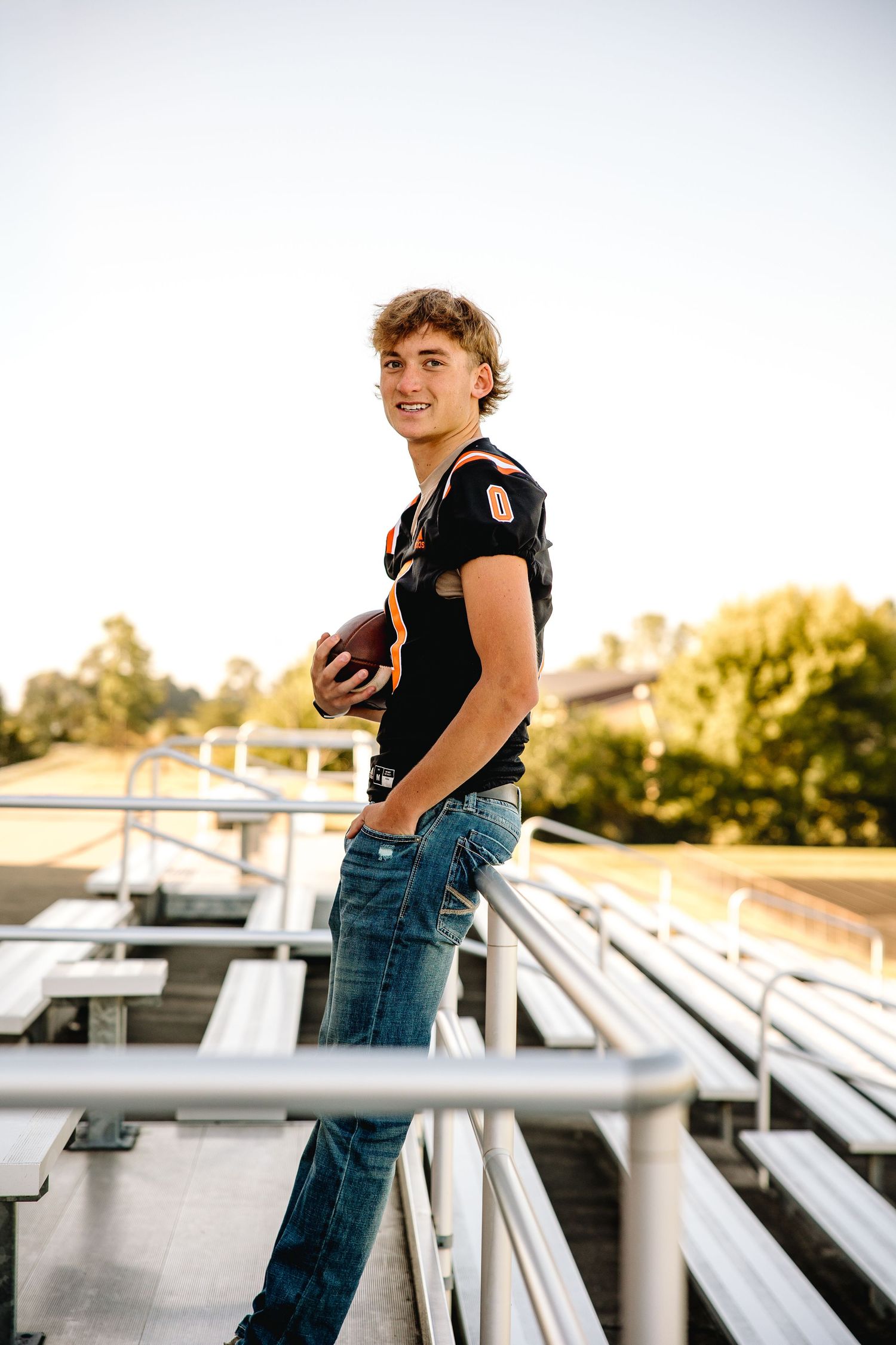 A smiling teenage boy in a black football jersey holds a football while standing on bleachers in jeans outdoors.