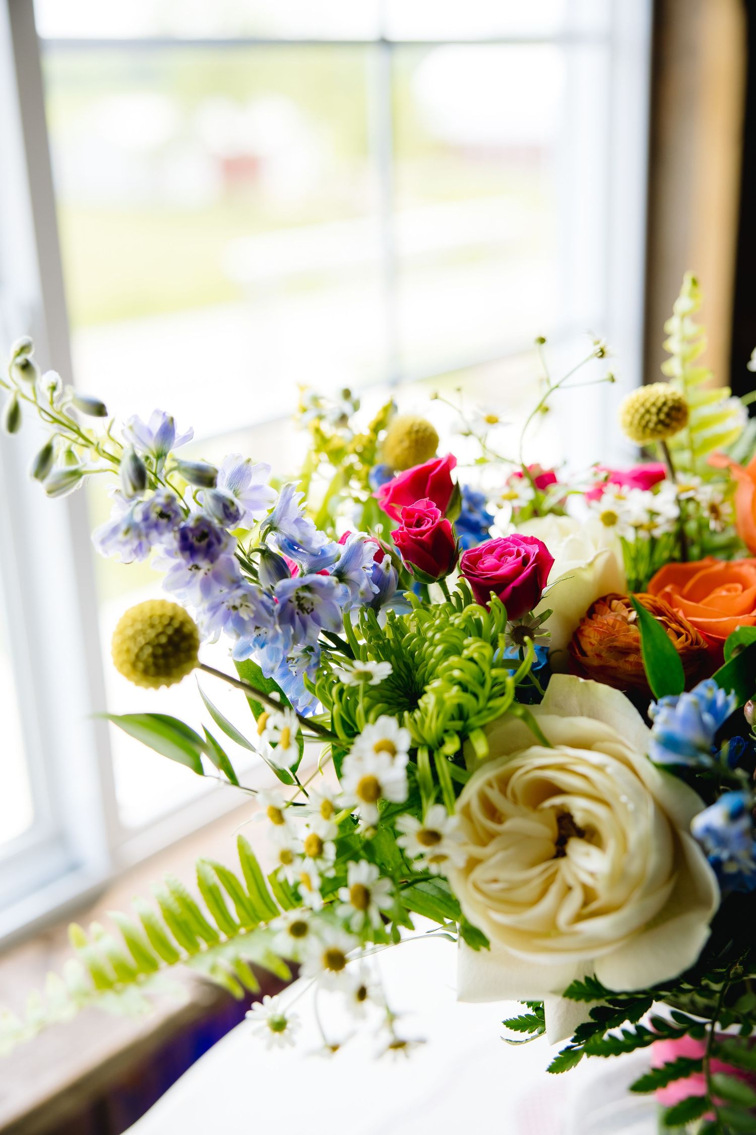 Colorful wedding bouquet with white roses, pink blooms, orange flowers, daisies, and greenery arranged near a bright window.