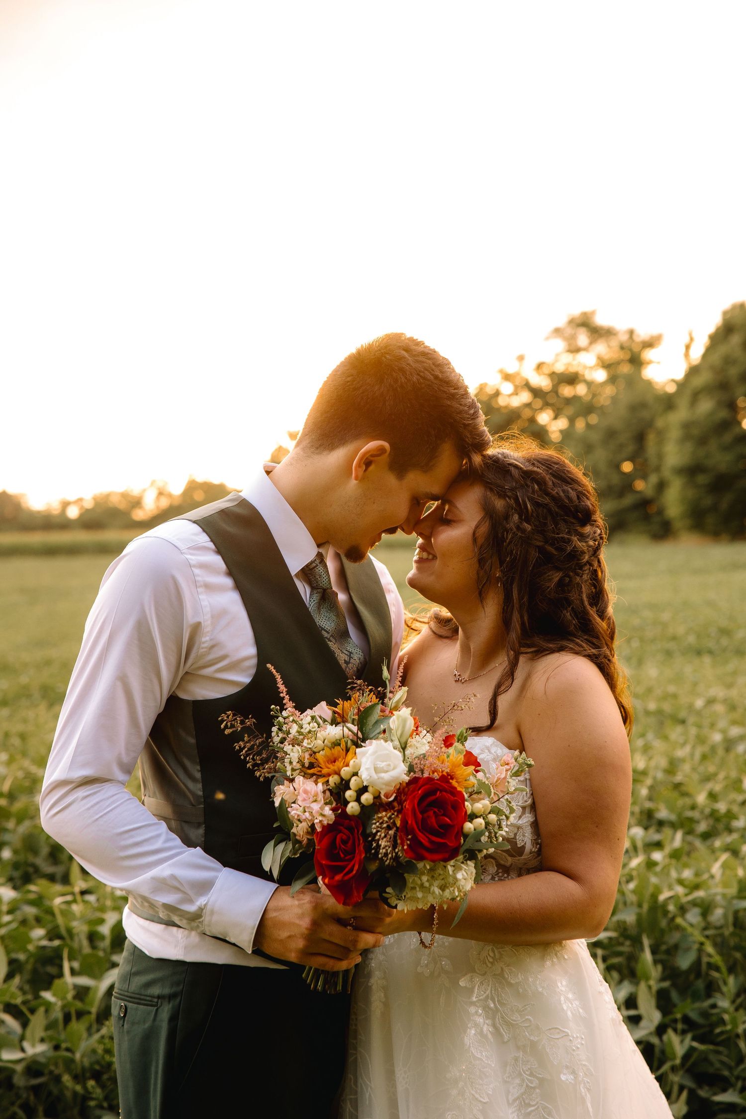 Bride and groom share a romantic golden hour moment in a field, holding a bouquet of red and white roses.