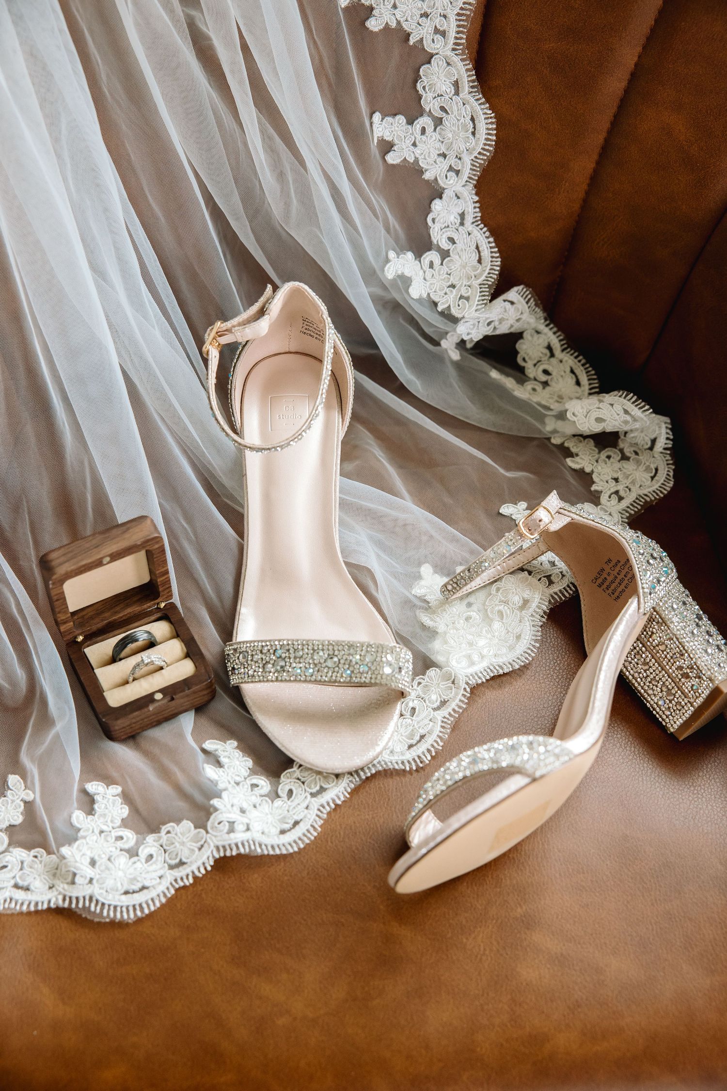 Elegant bridal heels with crystal embellishments displayed beside a lace wedding veil and wooden ring box on leather chair.