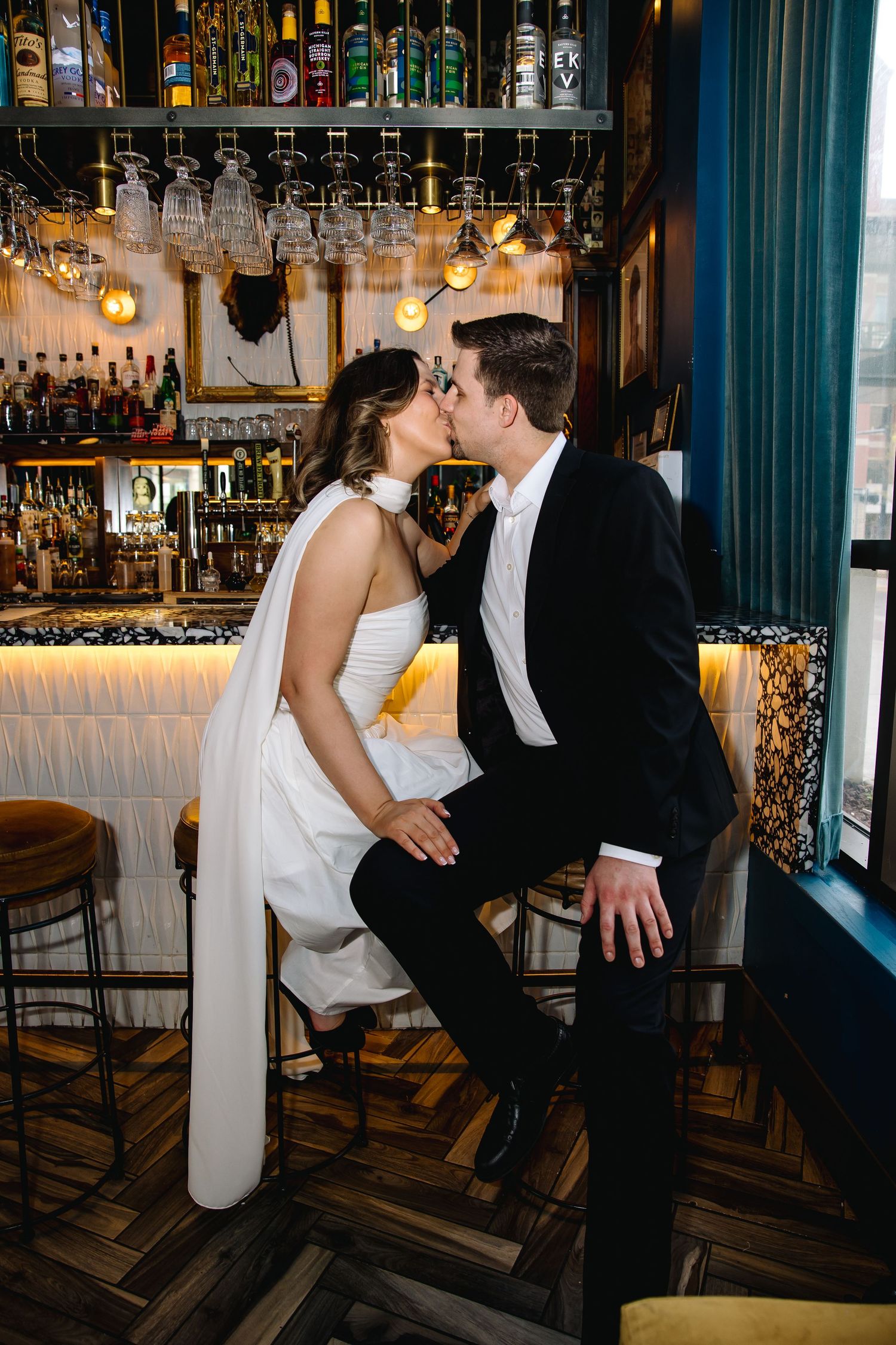 Bride and groom sharing a romantic kiss while seated at a stylish bar with bottles and hanging glasses.