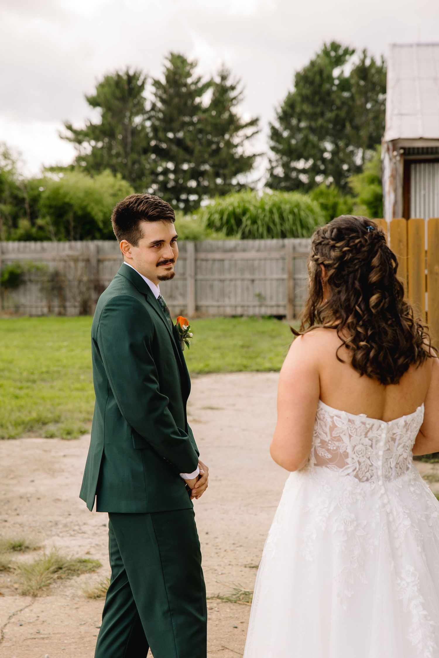 Groom in green suit smiling at bride in lace wedding dress during outdoor first look ceremony.