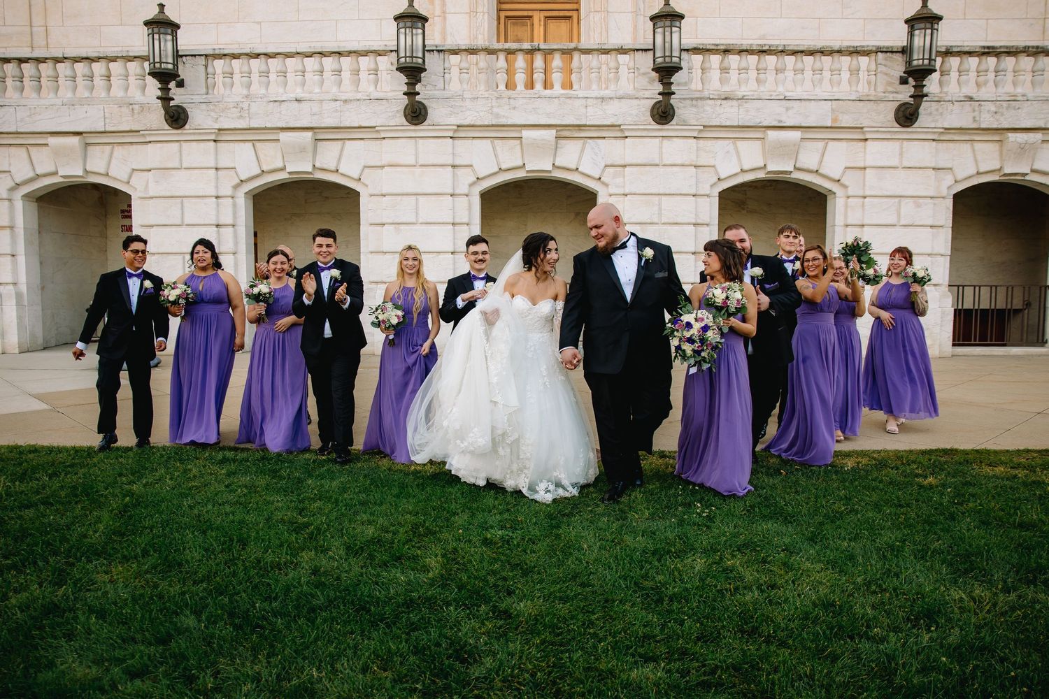 Bride and groom walk with wedding party in purple bridesmaid dresses outside elegant stone building.