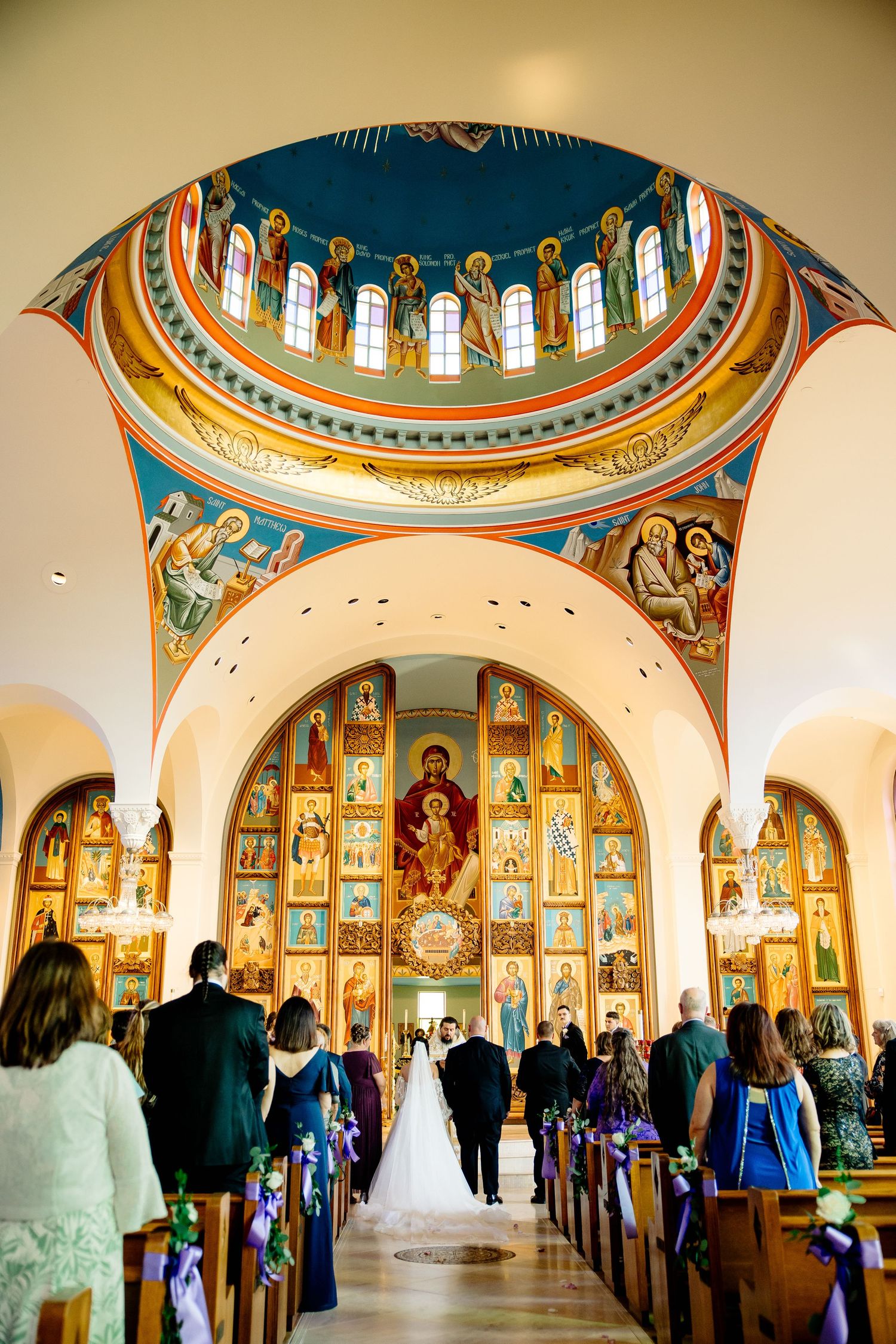 Bride walks down the aisle of an ornate Orthodox church with colorful icons and a stunning blue dome during wedding ceremony.