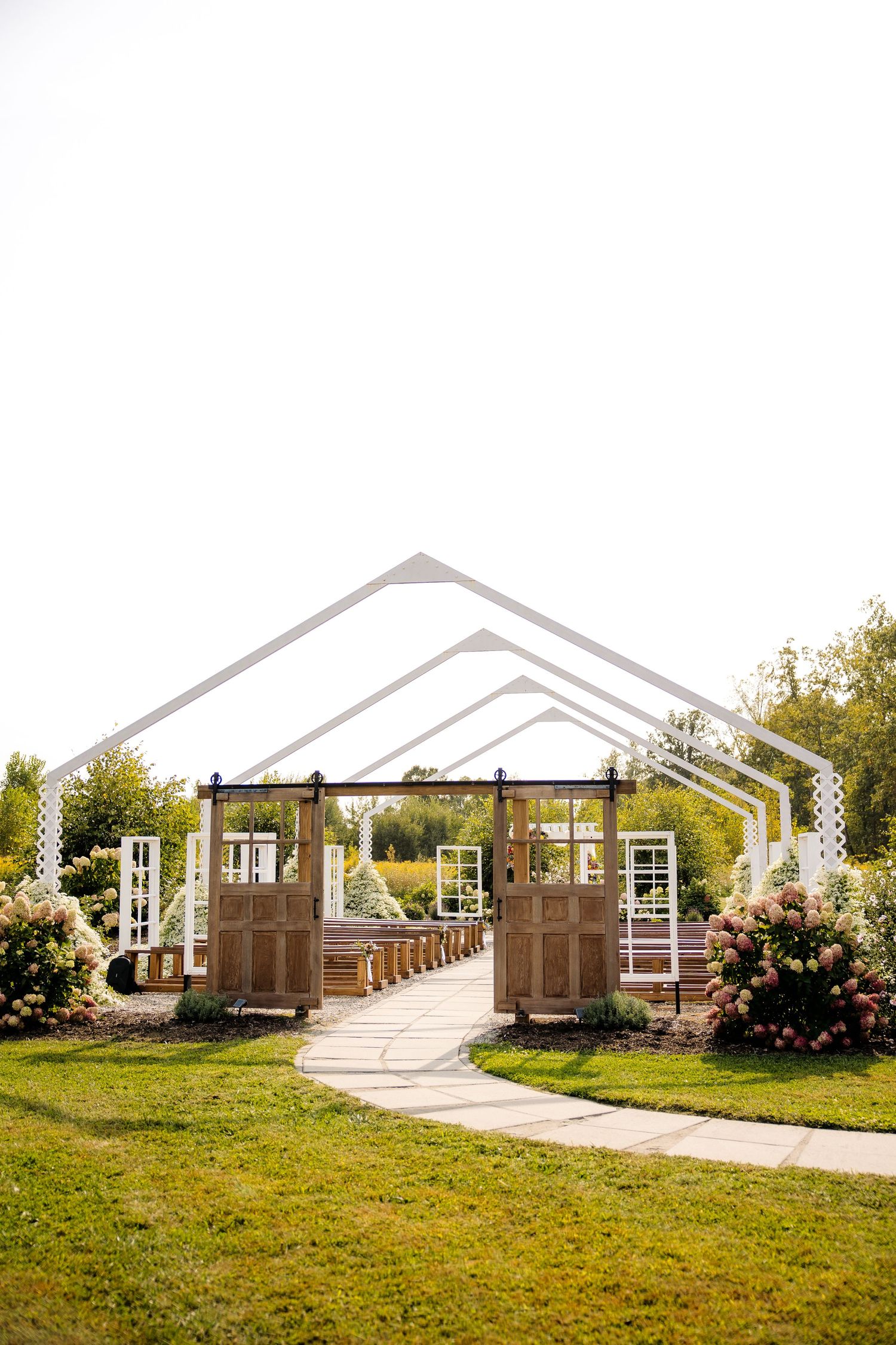 Outdoor wedding ceremony venue with white arch frames, wooden doors, stone pathway, and blooming pink floral bushes on green lawn.