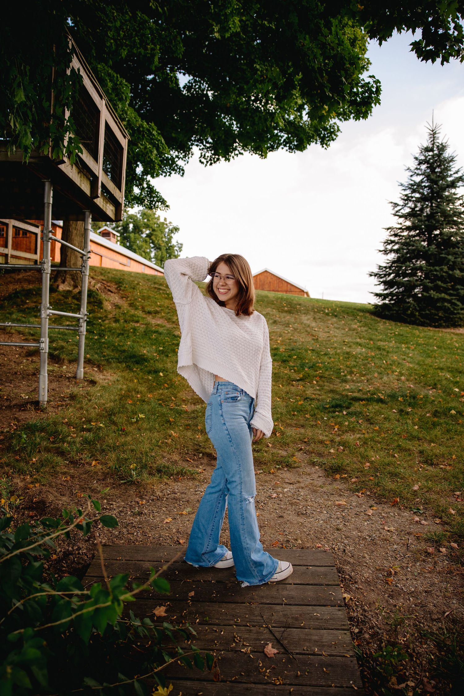 Young woman in white sweater and flared jeans smiles on wooden path surrounded by green trees and rustic barn.